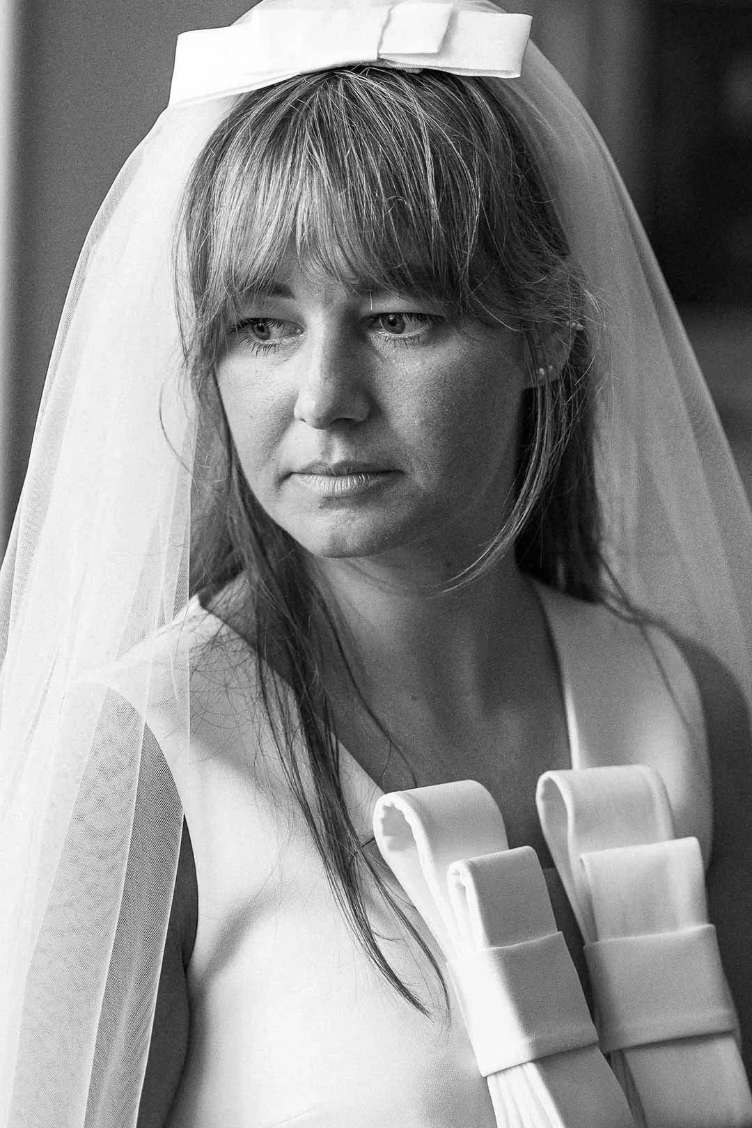 Zwarte en witte foto van een vrouw met een witte bruidshoed en een witte jurk, met een serieuze uitdrukking.