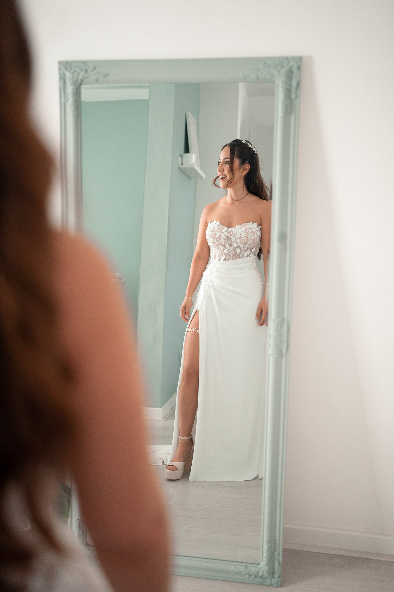 A woman in a wedding dress standing in front of a mirror, smiling, with a close-up of her reflection visible.