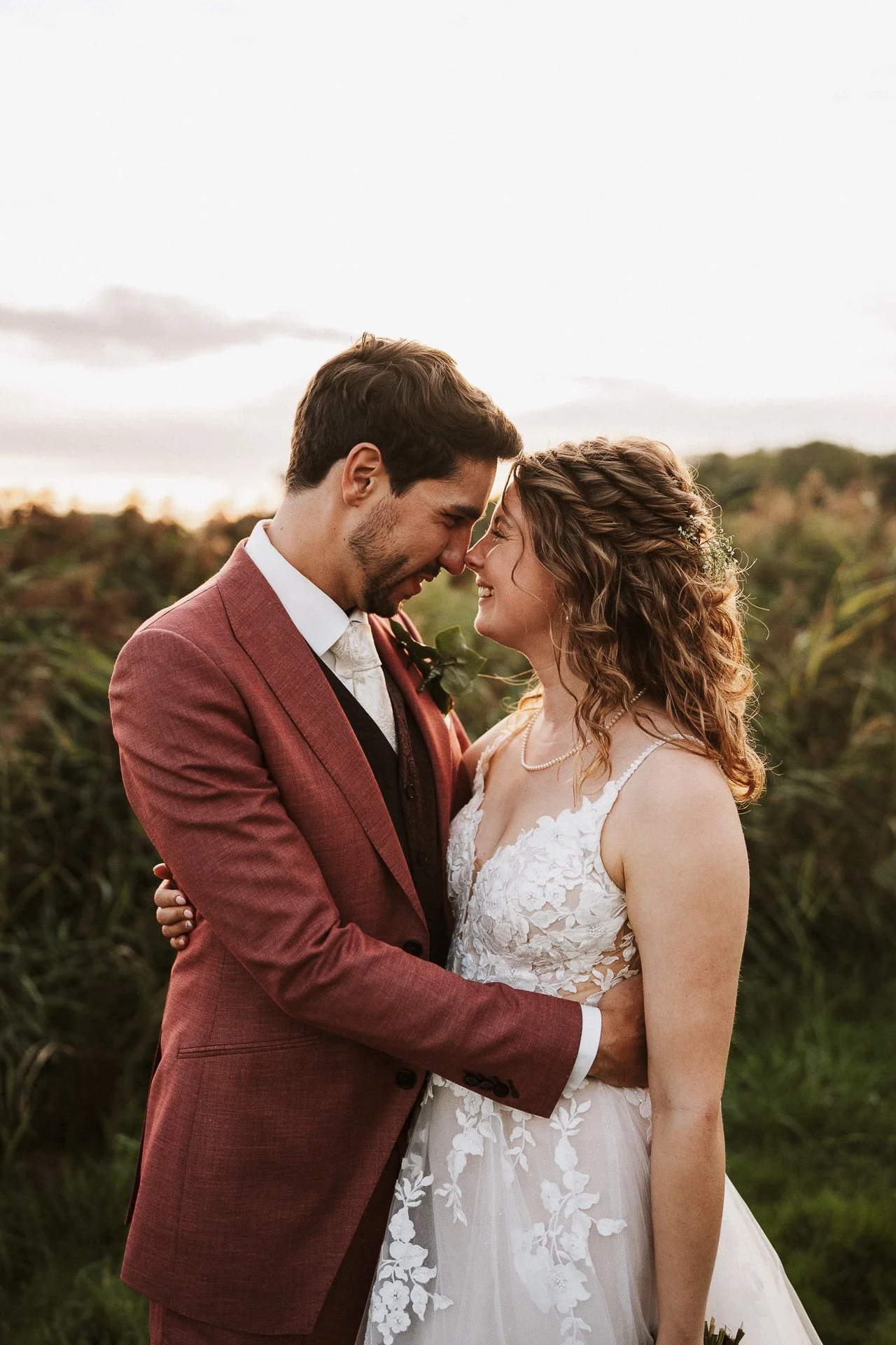 Twee mensen, een man in een bruinpak en een vrouw in een witte trouwjurk, staan dicht bij elkaar met voorhoofd tegen voorhoofd op een veld tijdens zonsondergang.