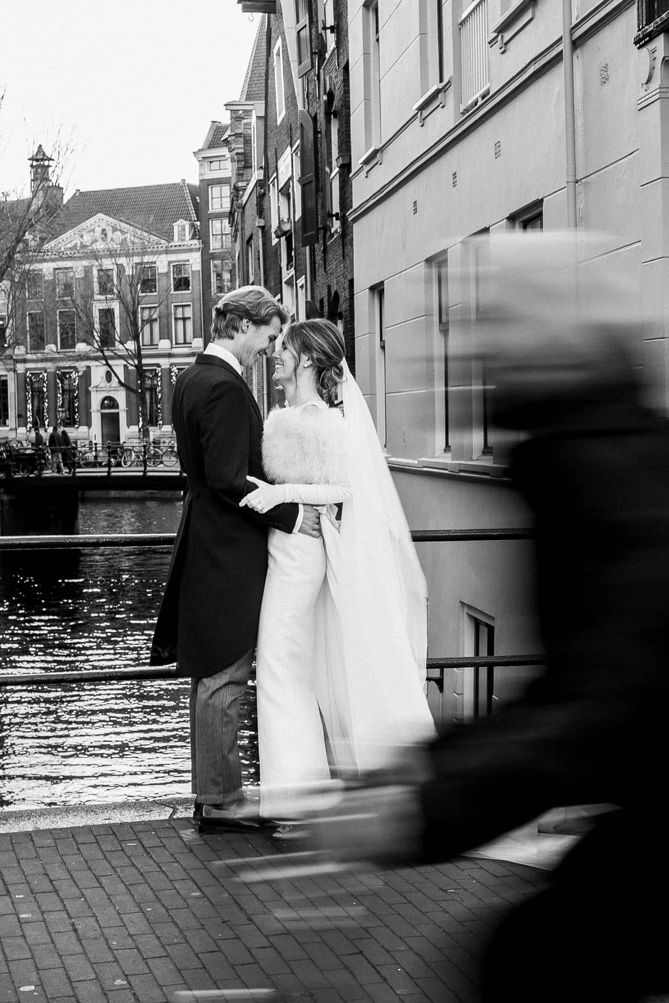 A black and white photo of a bride and groom standing closely on a canal side, smiling at each other, with buildings and trees in the background. There is movement blur of a passing boat in the foreground.