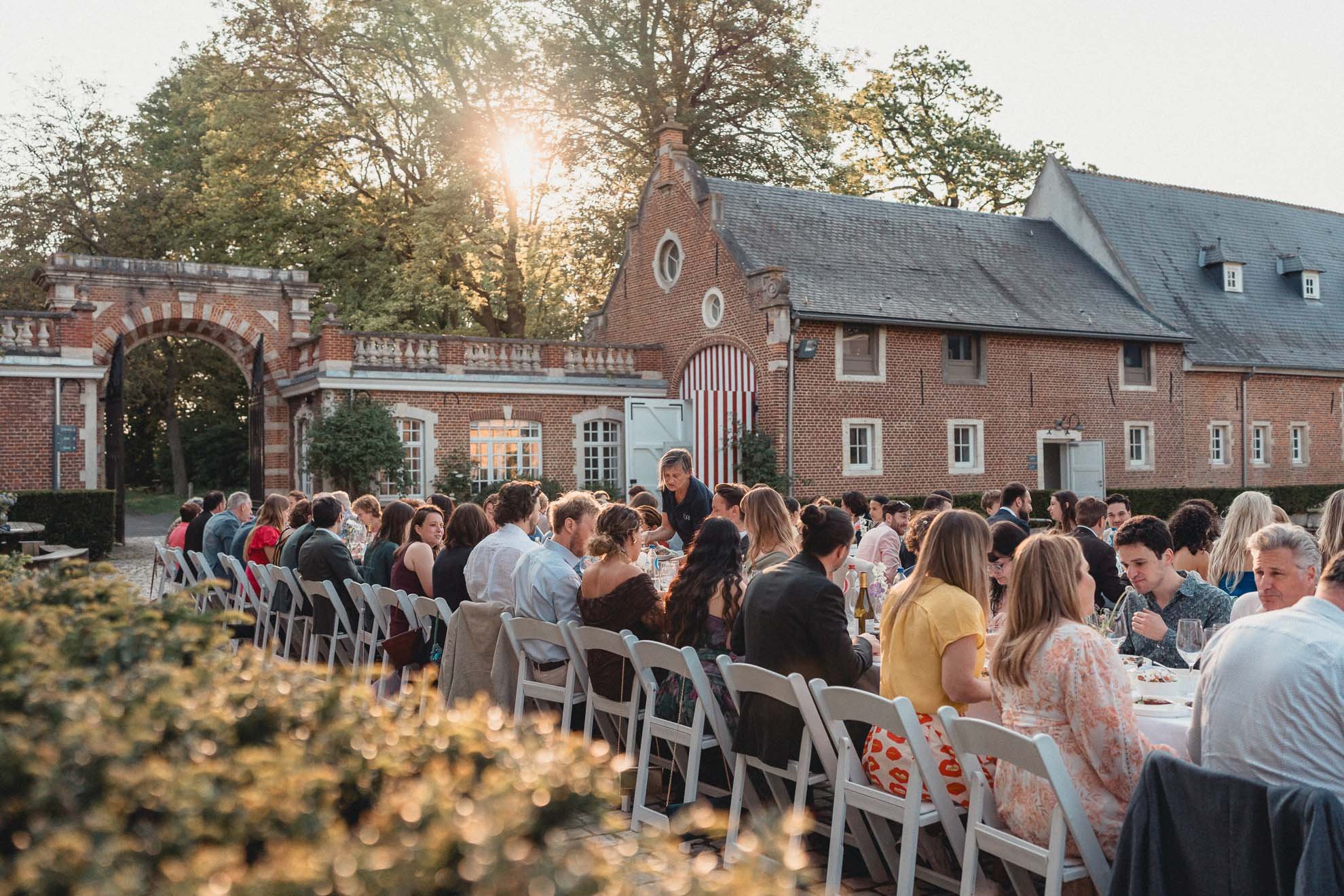An outdoor event with a large group of people seated at long tables in front of a historic brick building with a red and white striped door. The setting sun shines through trees in the background.