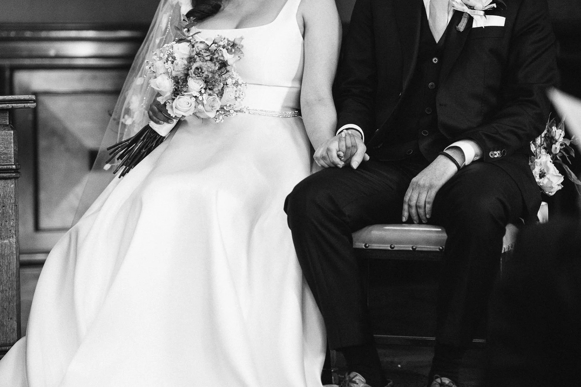 Black and white photo of a bride and groom holding hands during a wedding ceremony, sitting on a bench in a church, the bride in a white gown with a bouquet of flowers, and the groom in a dark suit.