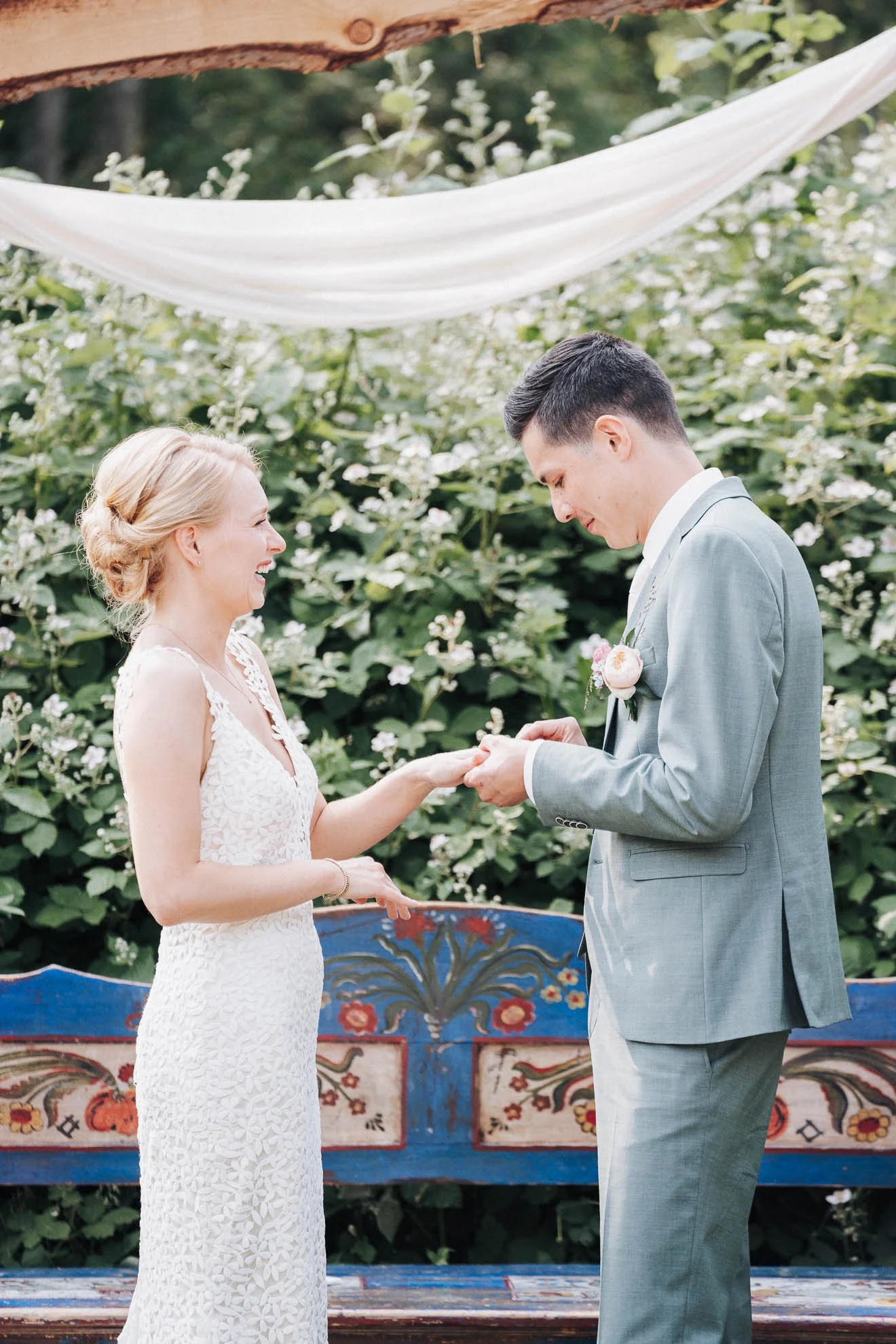 A bride and groom exchange wedding vows outdoors, standing in front of a lush green background with white flowers and a painted bench indicates a romantic setting.