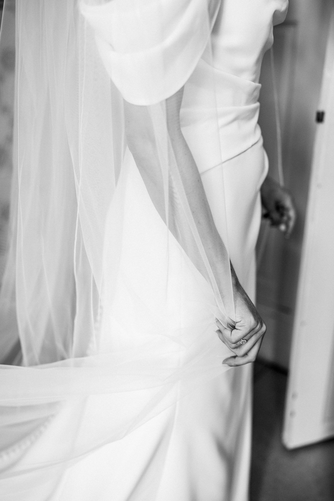 A woman in a wedding dress is sitting with her hand resting on her lap, showing a wedding ring. She is also wearing a veil.