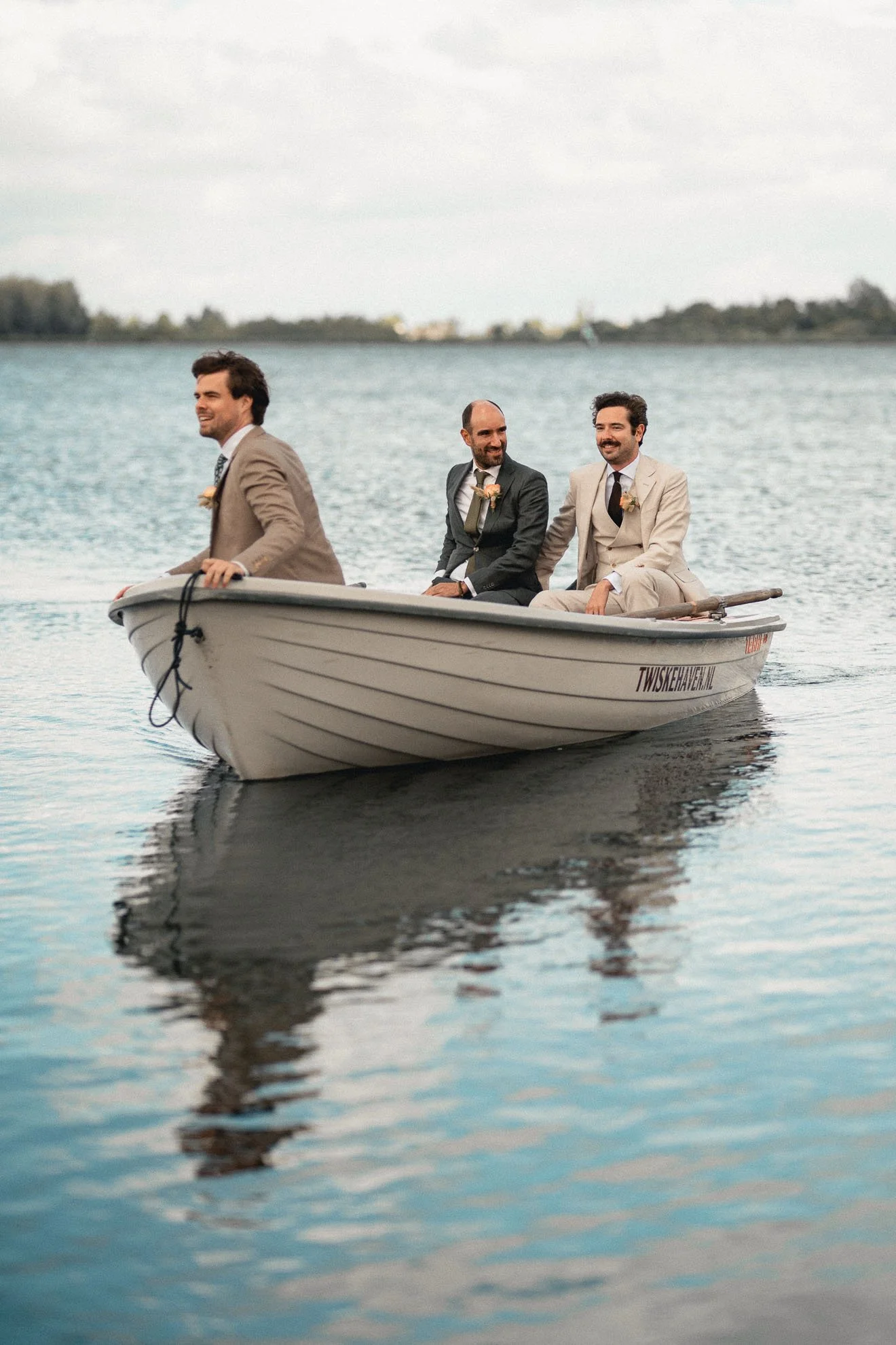 Three men in suits are sitting in a small boat on a body of water, smiling and looking to the side, with trees and a cloudy sky in the background.