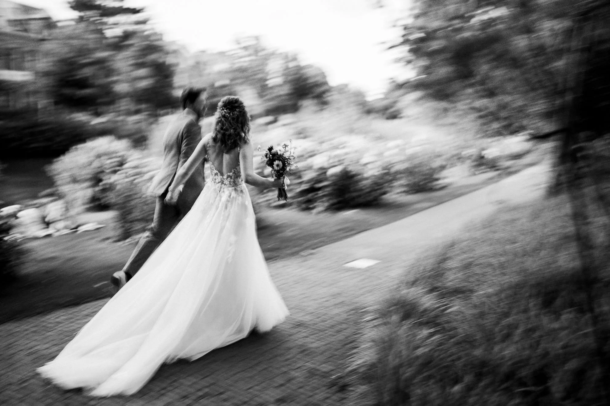 A black and white photograph of a bride and groom walking hand in hand outdoors, with the bride holding a bouquet of flowers and wearing a long, flowing wedding dress.