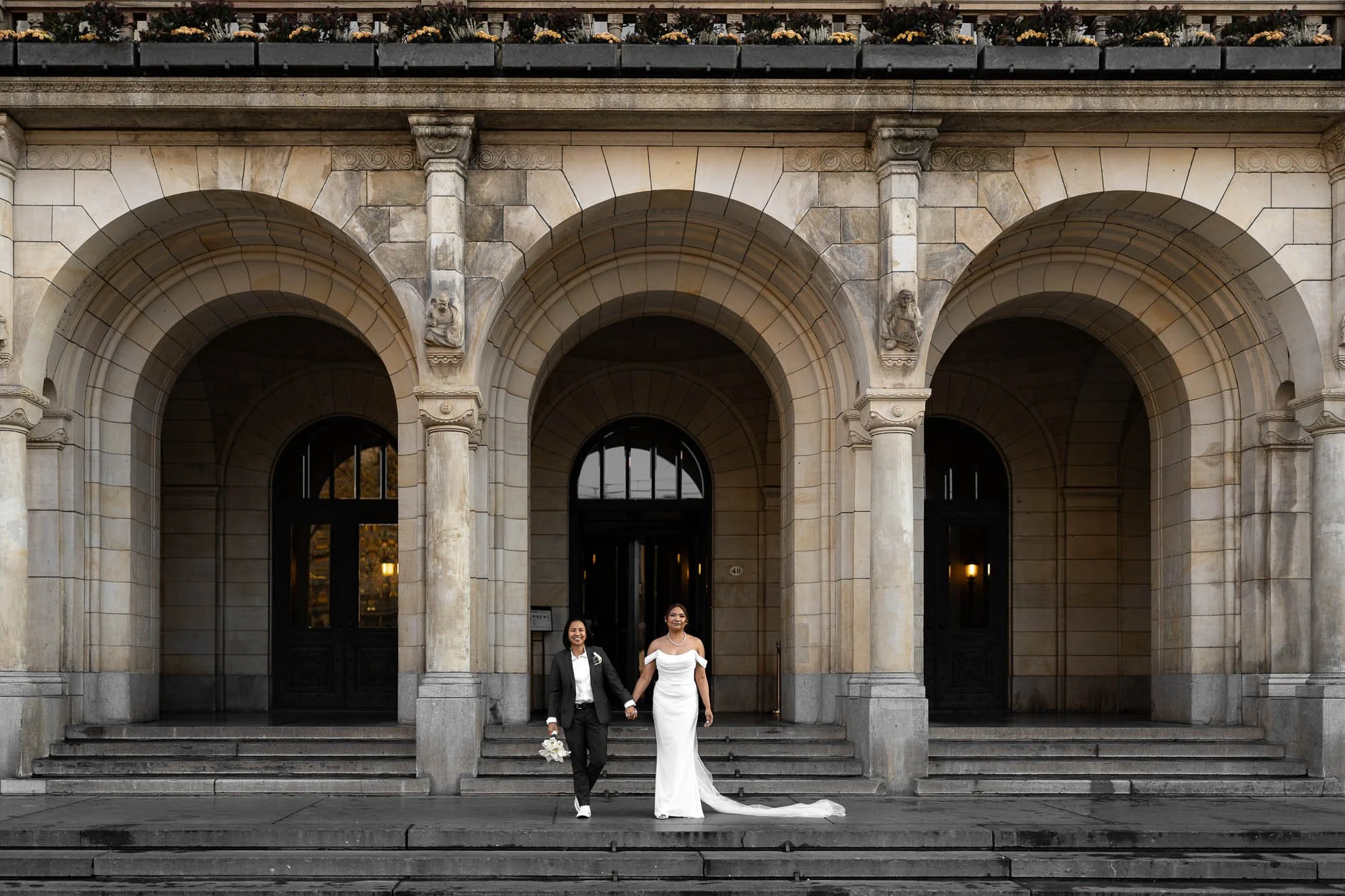 A wedding couple standing on steps in front of an ornate stone building with three archways. The groom is wearing a black suit and white shirt, holding a bouquet, and the bride is in a white wedding dress with short sleeves, holding hands with the gr