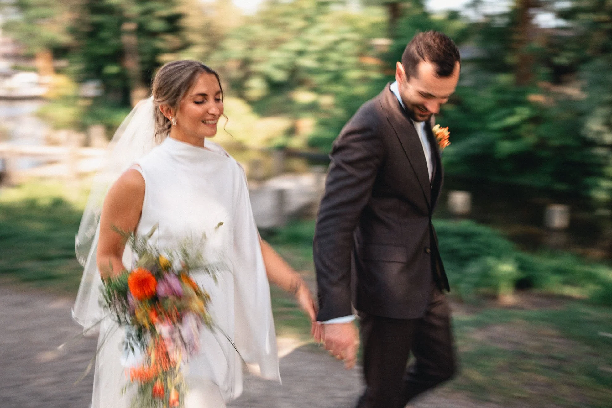 A bride and groom walking hand in hand outdoors, smiling, with the bride holding a colorful bouquet of flowers, greenery in the background.