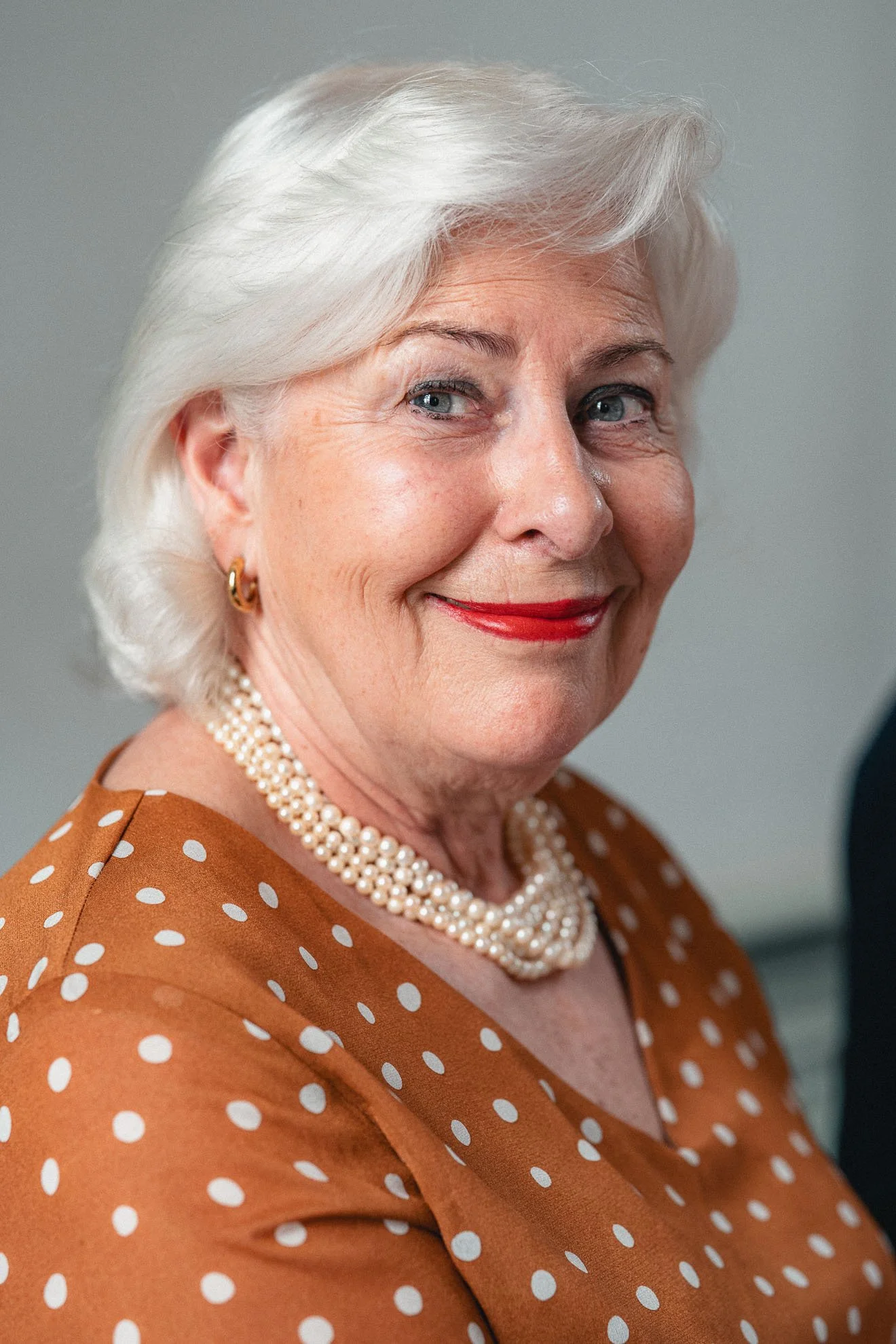 Close-up of a smiling older woman with white hair, blue eyes, wearing red lipstick, pearl necklace, gold earrings, and a brown dress with white polka dots.