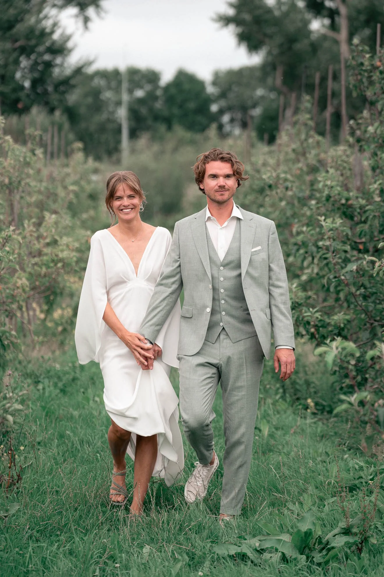 A smiling woman in a white dress and a man in a gray suit walk hand-in-hand through a green orchard.