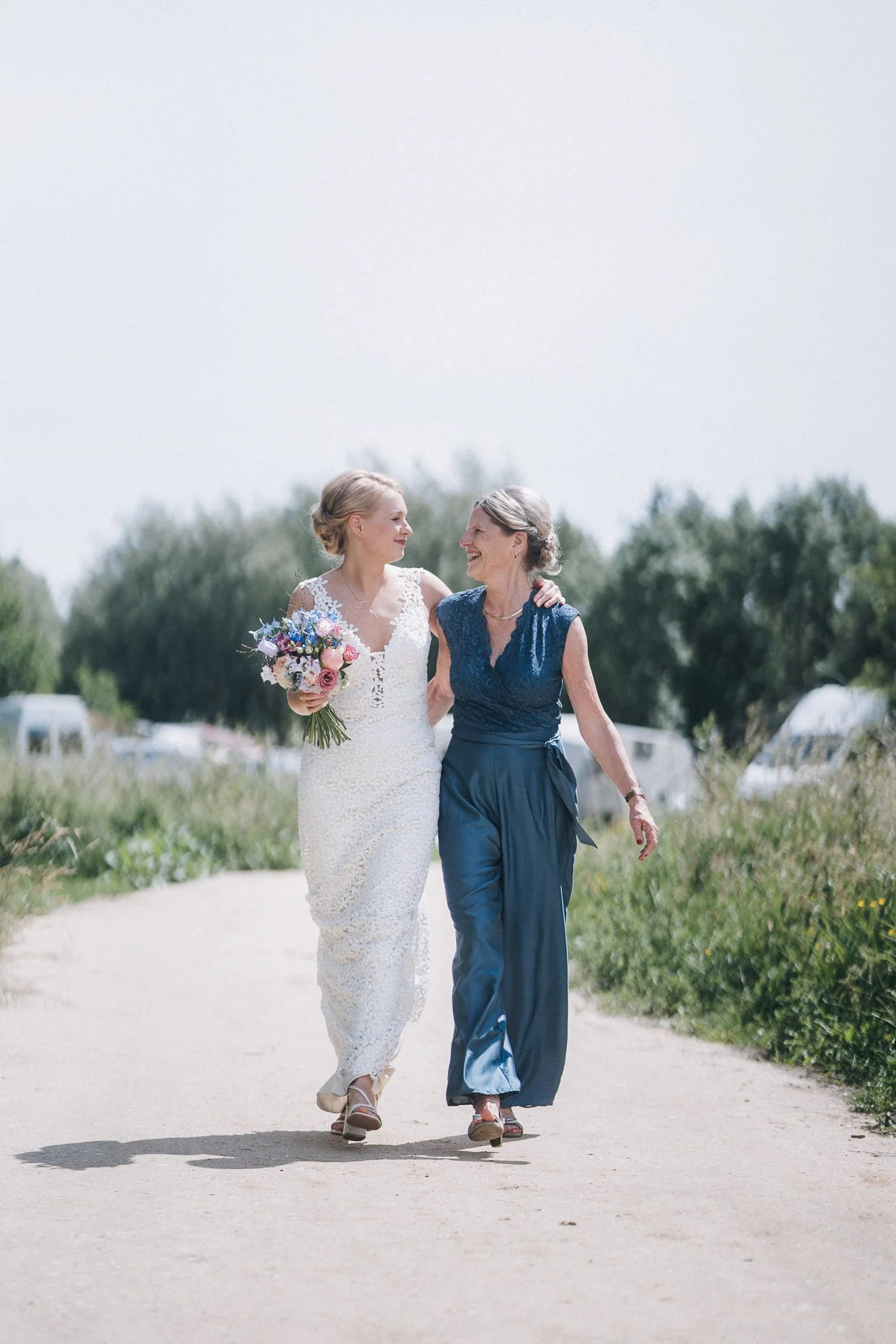 A bride in a white lace dress holding a bouquet of pink and purple flowers, walking arm-in-arm with an older woman in a blue lace top and matching pants on a sunny day outdoors.