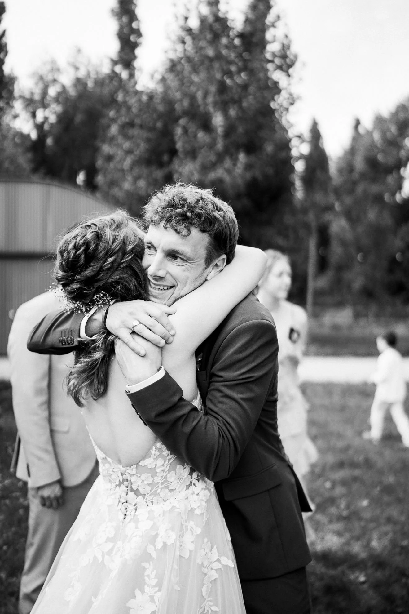 A bride and groom are hugging during a wedding celebration outdoors. The groom is smiling and dressed in a dark suit, while the bride is in a lace wedding dress. There are blurred wedding guests and trees in the background.