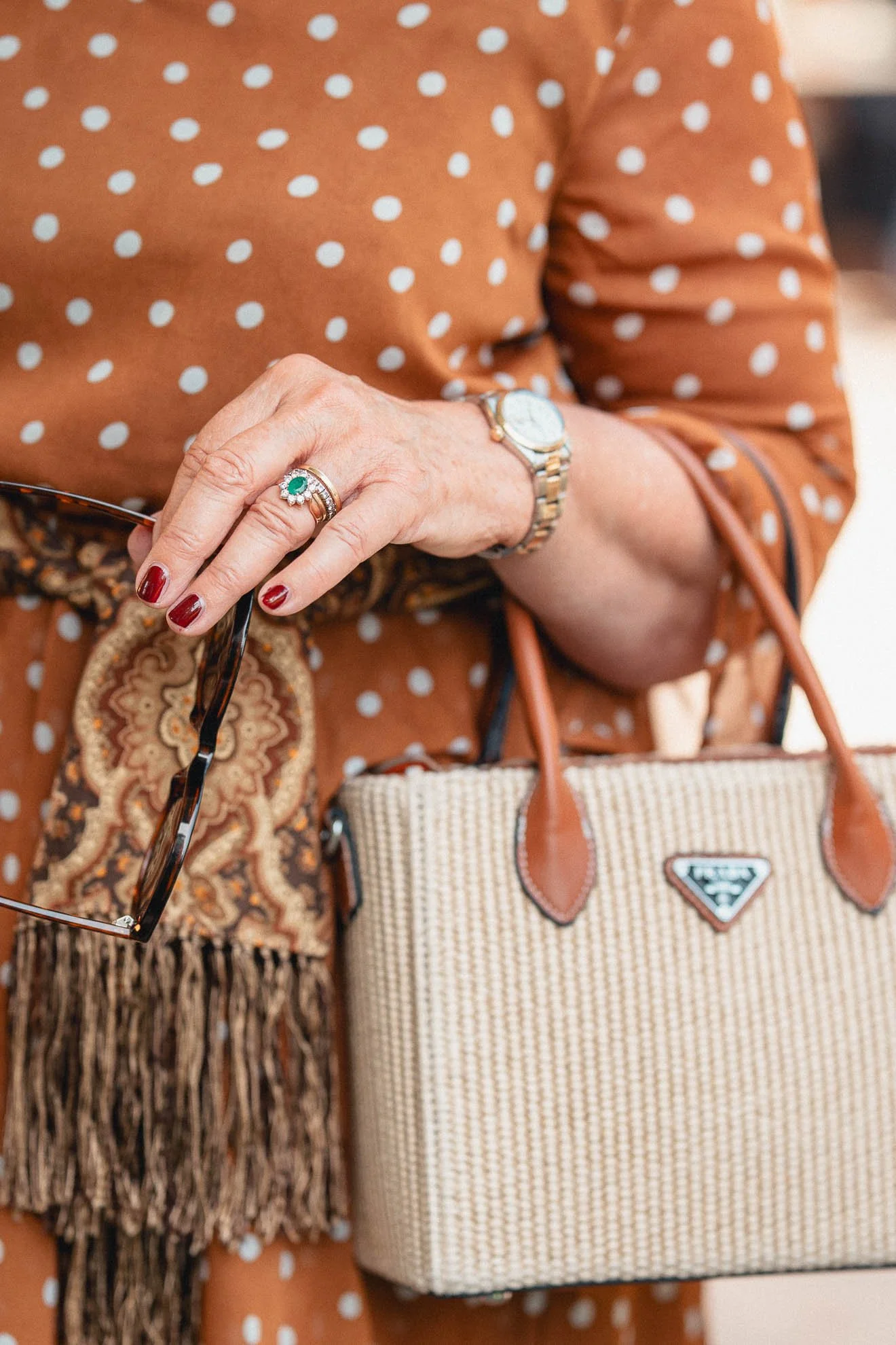 Close-up of a woman wearing a polka-dot dress, holding sunglasses, with red painted nails, a floral ring, a watch, and a Prada bag visible in the frame.
