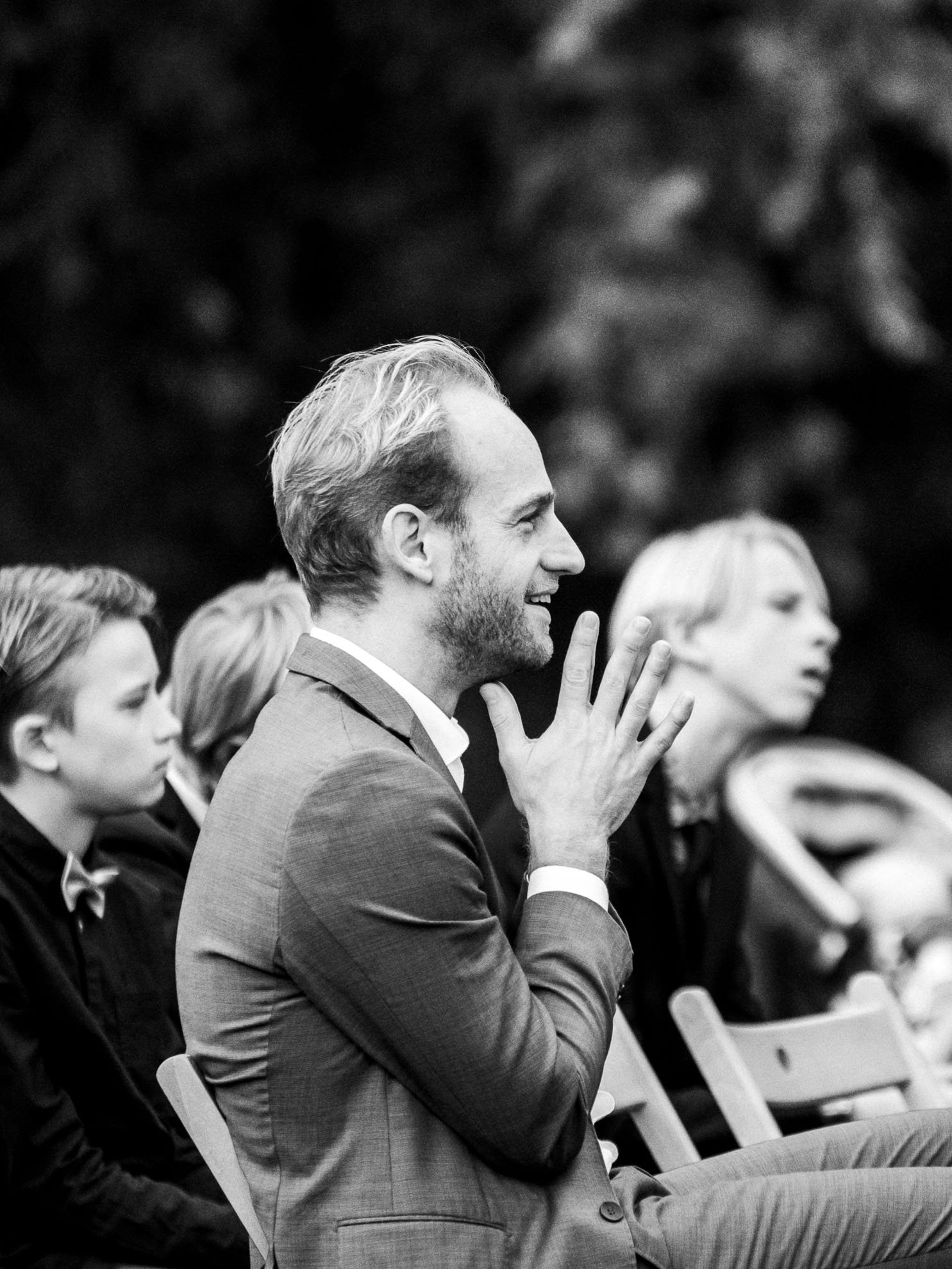 A man in a suit with light-colored hair, smiling and clapping, sitting in an outdoor setting with other well-dressed people.
