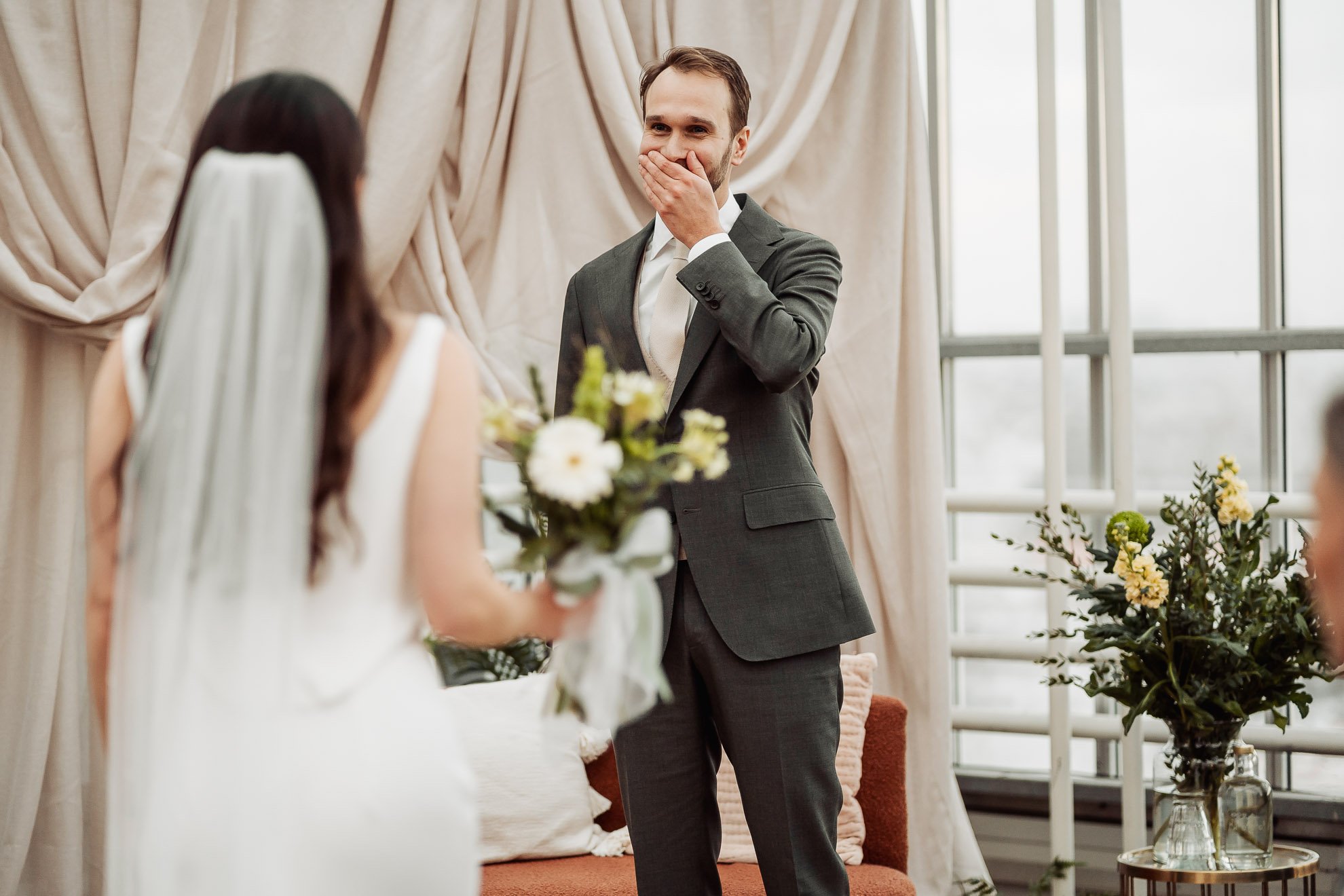 A man in a gray suit covering his mouth with his hand, smiling, during a wedding ceremony. A woman in a white dress holding a bouquet of flowers is in the foreground, with her back to the camera. The setting is decorated with curtains and flowers.