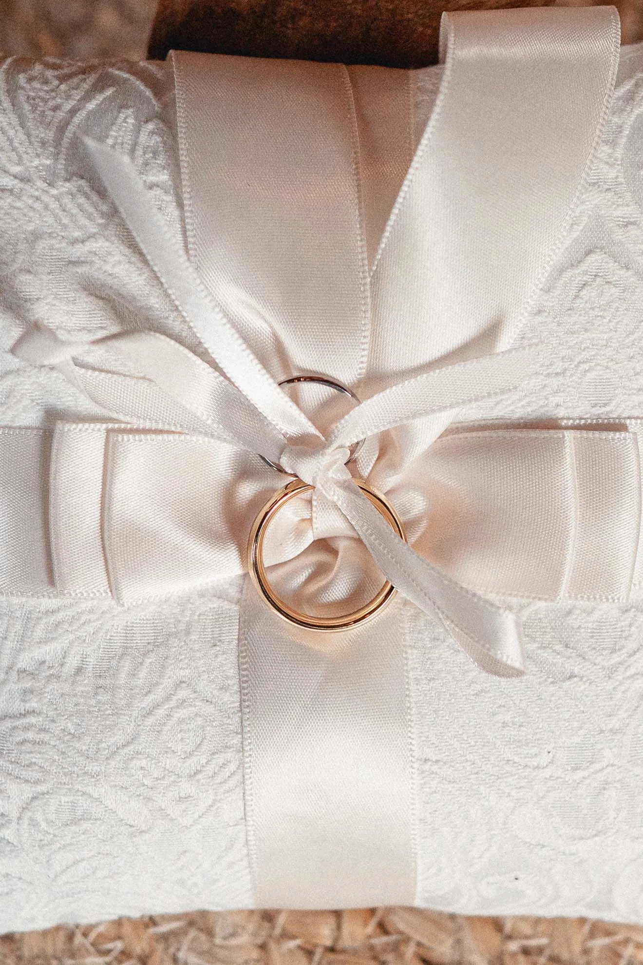 A close-up of a white wedding ring box with a ring inside, decorated with a satin ribbon and lace fabric, placed on a textured surface.