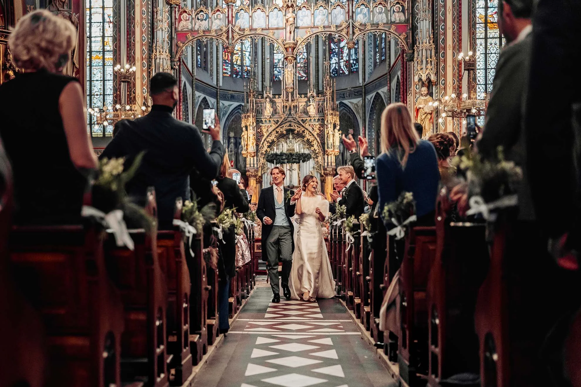 A newlywed couple walks down the aisle of a decorated church, smiling and holding hands, with guests on both sides taking pictures. The church has ornate stained glass windows and detailed religious artwork.
