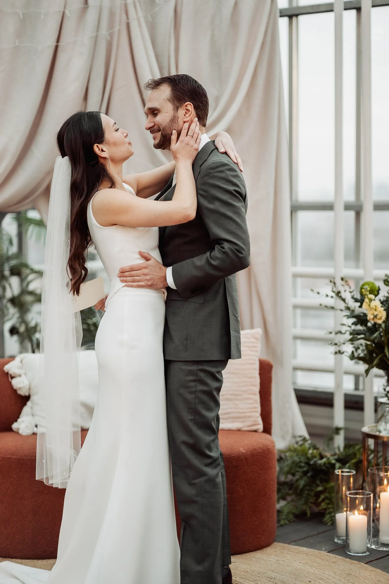 A bride and groom share a romantic moment indoors during their wedding, standing close with the bride touching the groom's face and the groom holding the bride's waist, with candles and plants in the background.