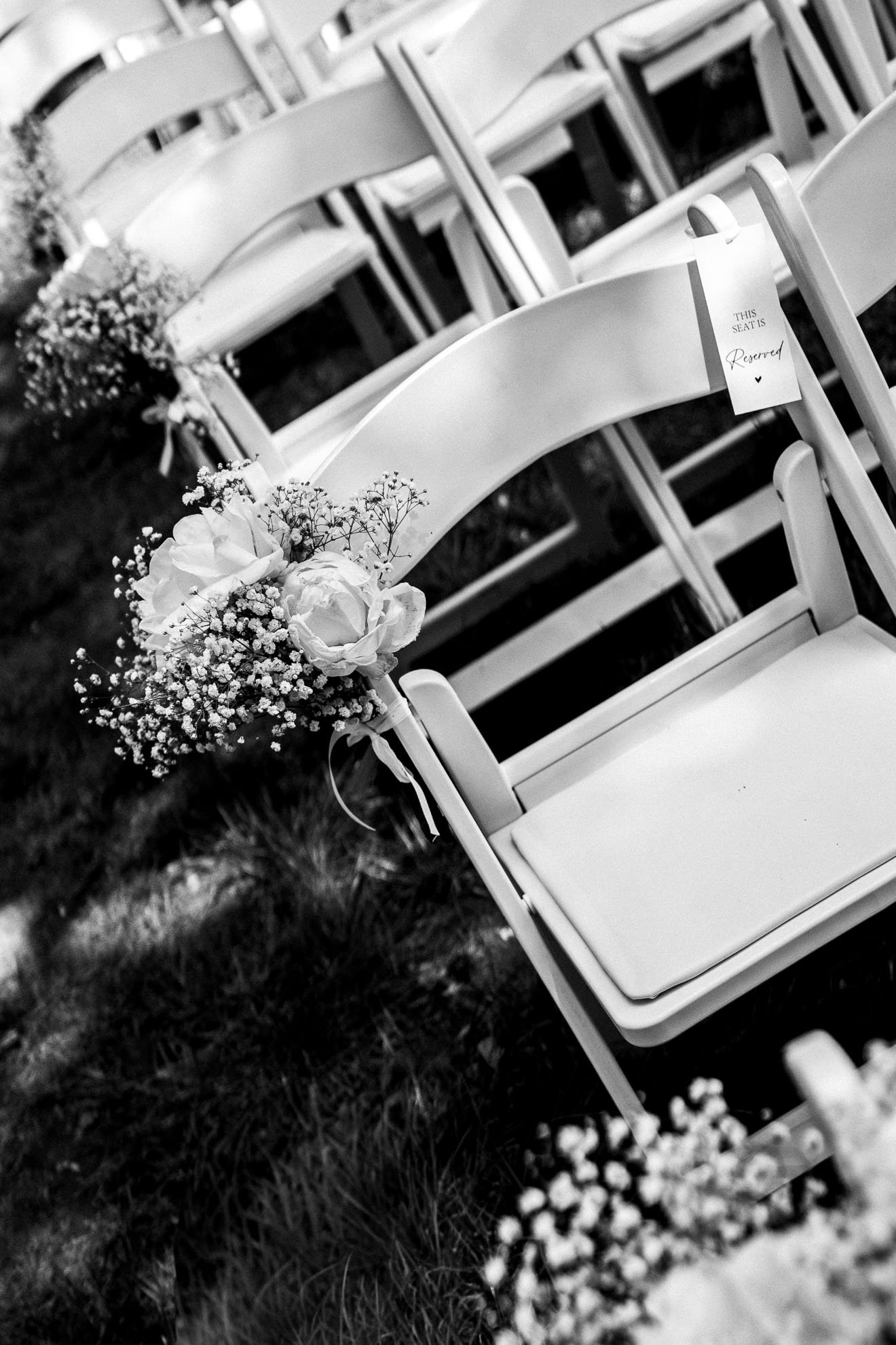 Rows of white chairs decorated with bouquets of flowers and reserved signs, set outside on grass for a wedding or event.