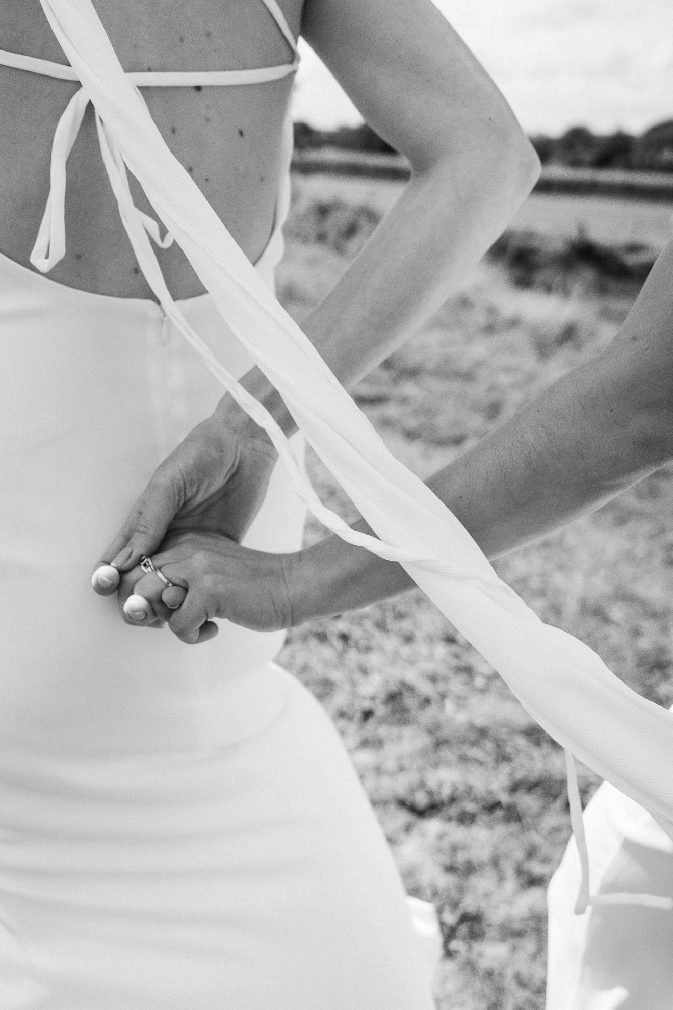 A close-up black-and-white photo of a woman wearing a sleeveless dress with tie straps, holding hands with someone, outdoors on a beach.