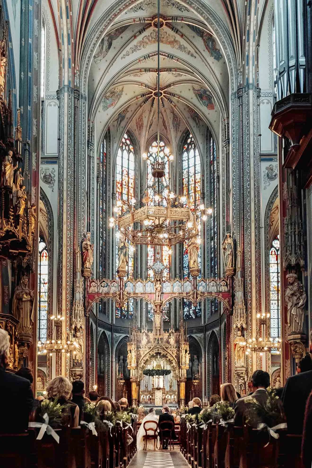 Interior view of a grand church or cathedral during a wedding ceremony, with people seated in pews, ornate decorations, stained glass windows, and chandeliers hanging from the vaulted ceiling.