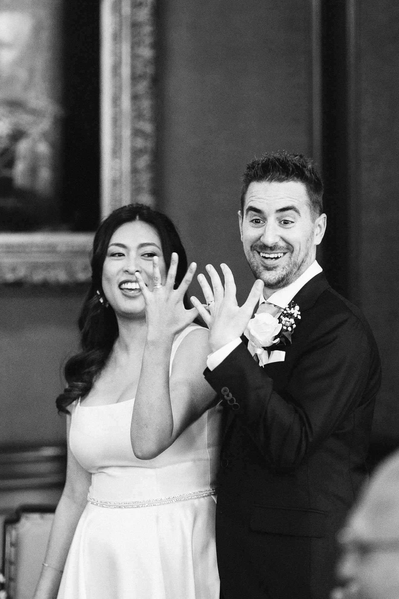 A black and white photo of a bride and groom at a wedding reception. The bride is wearing a sleeveless white wedding dress with a beaded waist detail, and the groom is in a black tuxedo with a boutonniere. They are showing their rings on their left h