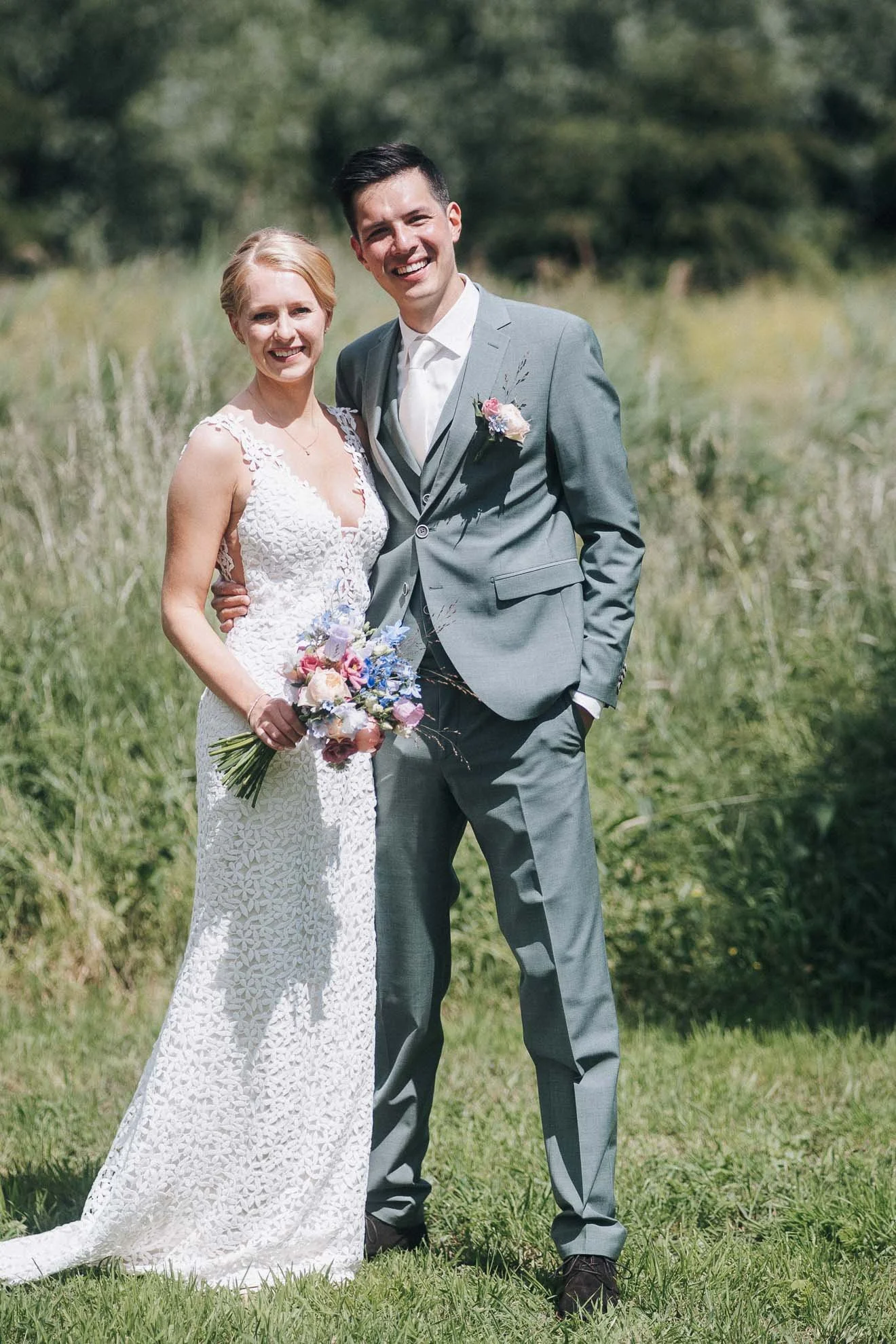 A happy couple in wedding attire standing outdoors in a grassy field, with the woman holding a bouquet of flowers and both smiling at the camera.