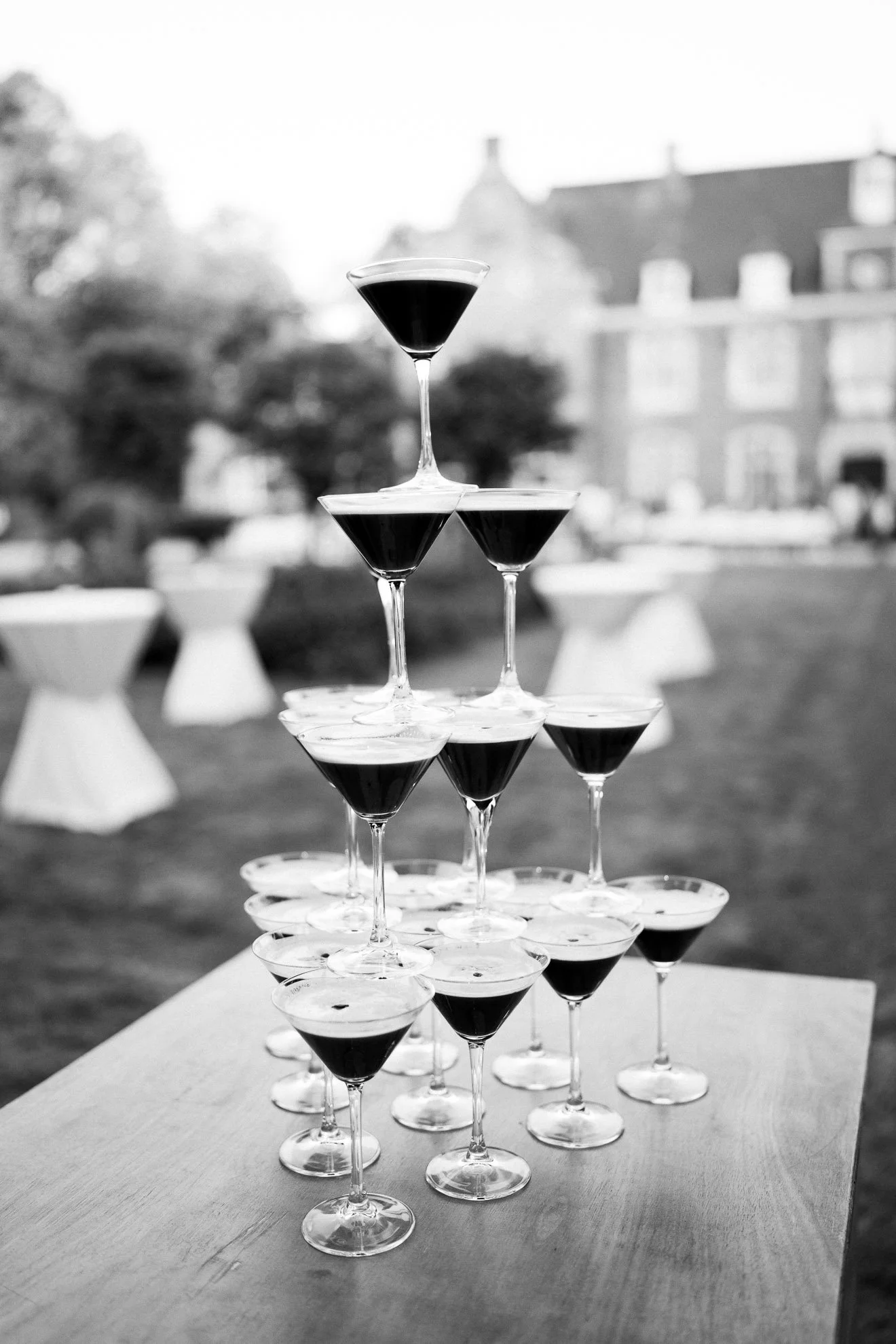 A pyramid of stacked martini glasses filled with dark liquid, set outdoors with trees and buildings in the background.