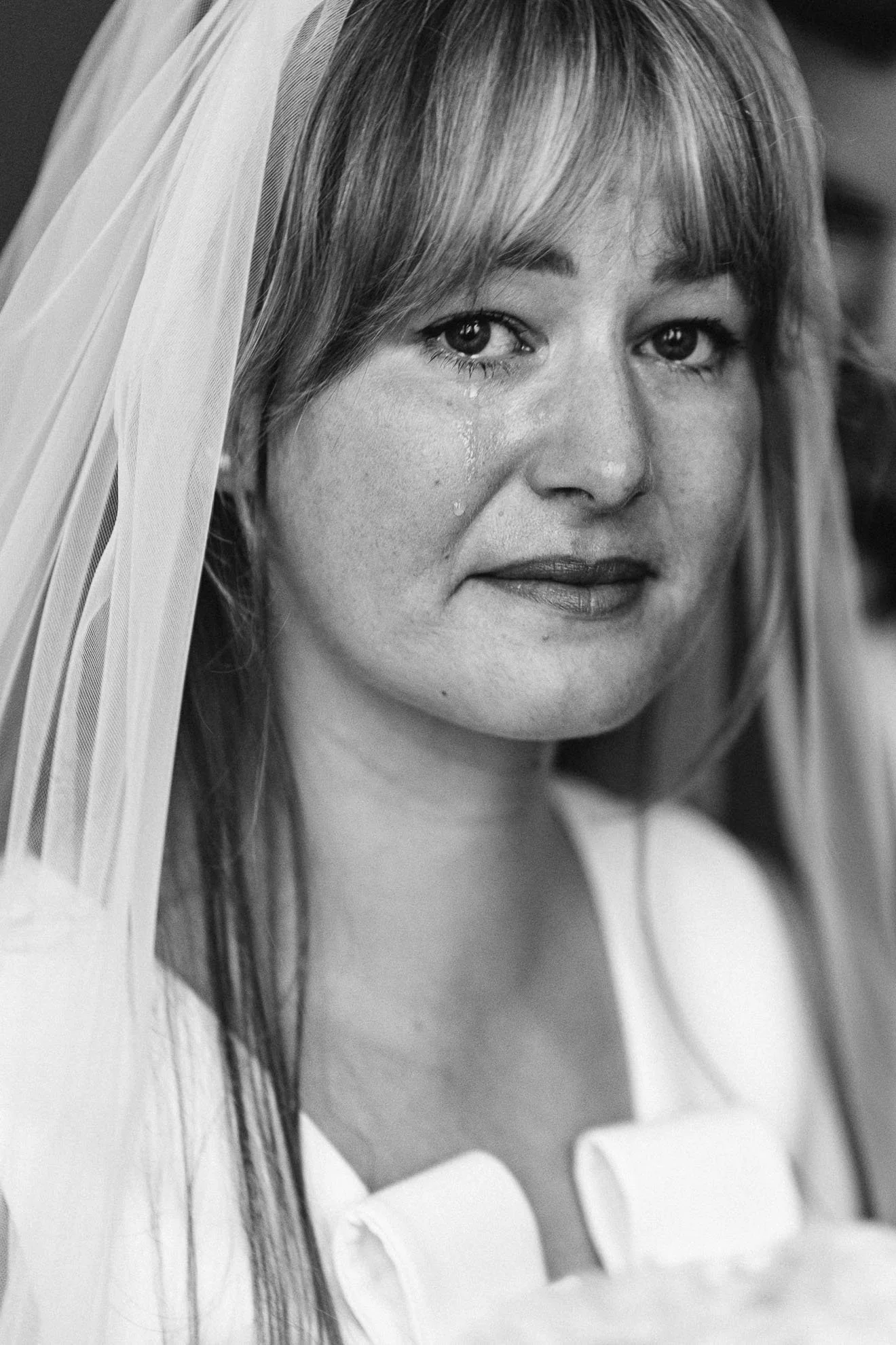 A black and white photograph of a woman in a wedding dress, with a veil partially covering her head, looking directly at the camera with tears on her face.