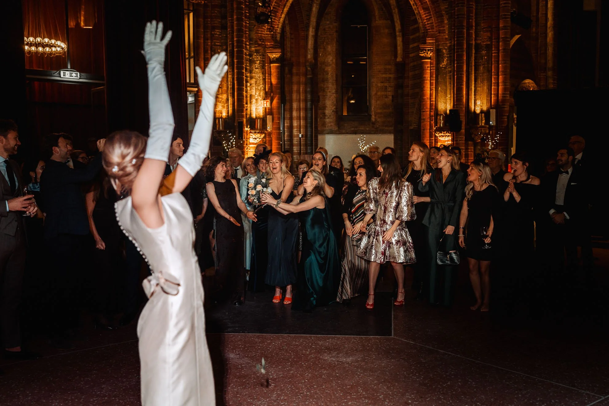 A woman in a white dress and long gloves raises her hands in front of a lively crowd at an indoor event with brick walls and warm lighting.