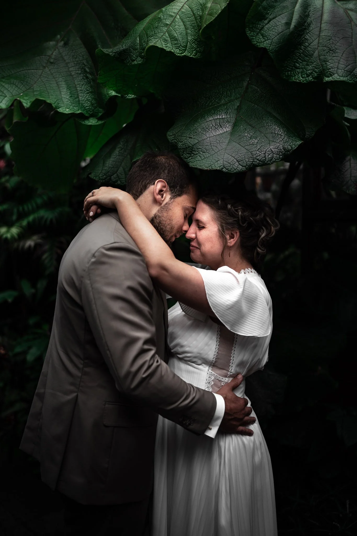 Een stel dat in een groene, natuurlijke omgeving dicht bij elkaar staat en elkaar liefkoost, met grote bladeren boven hen.
