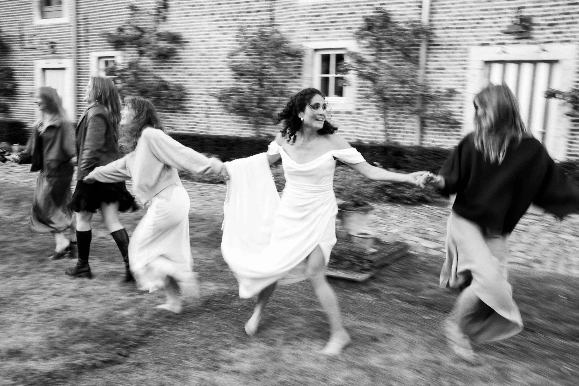 Black and white photo of women holding hands, dancing outdoors in front of a brick building.