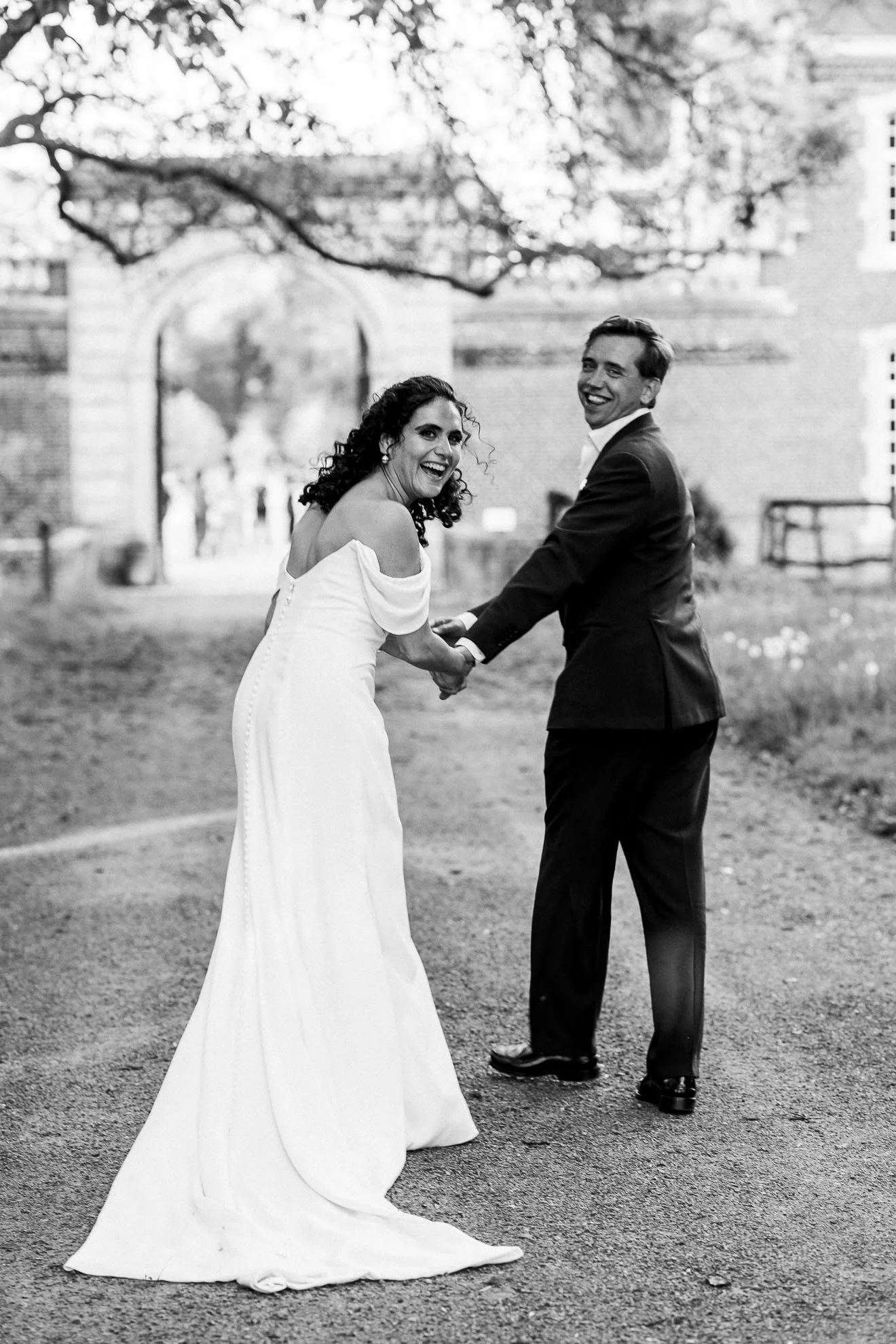 A happy bride and groom holding hands and smiling outdoors on a wedding day, with trees and a brick building in the background.