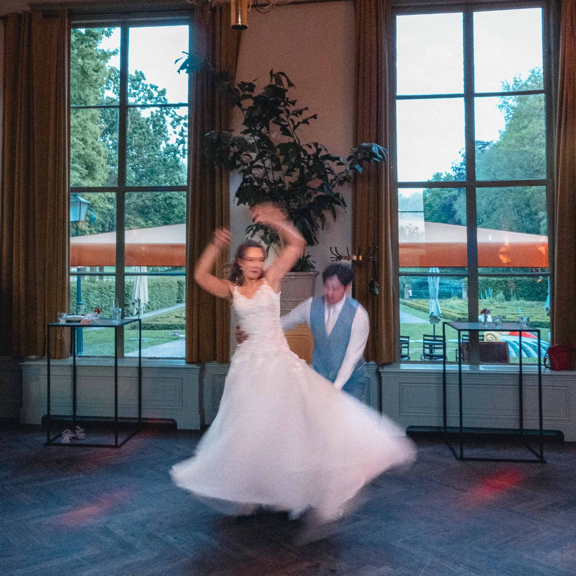 Two people dancing in front of large windows with a view of trees and a cloudy sky outside, in a room with dark flooring and curtains.