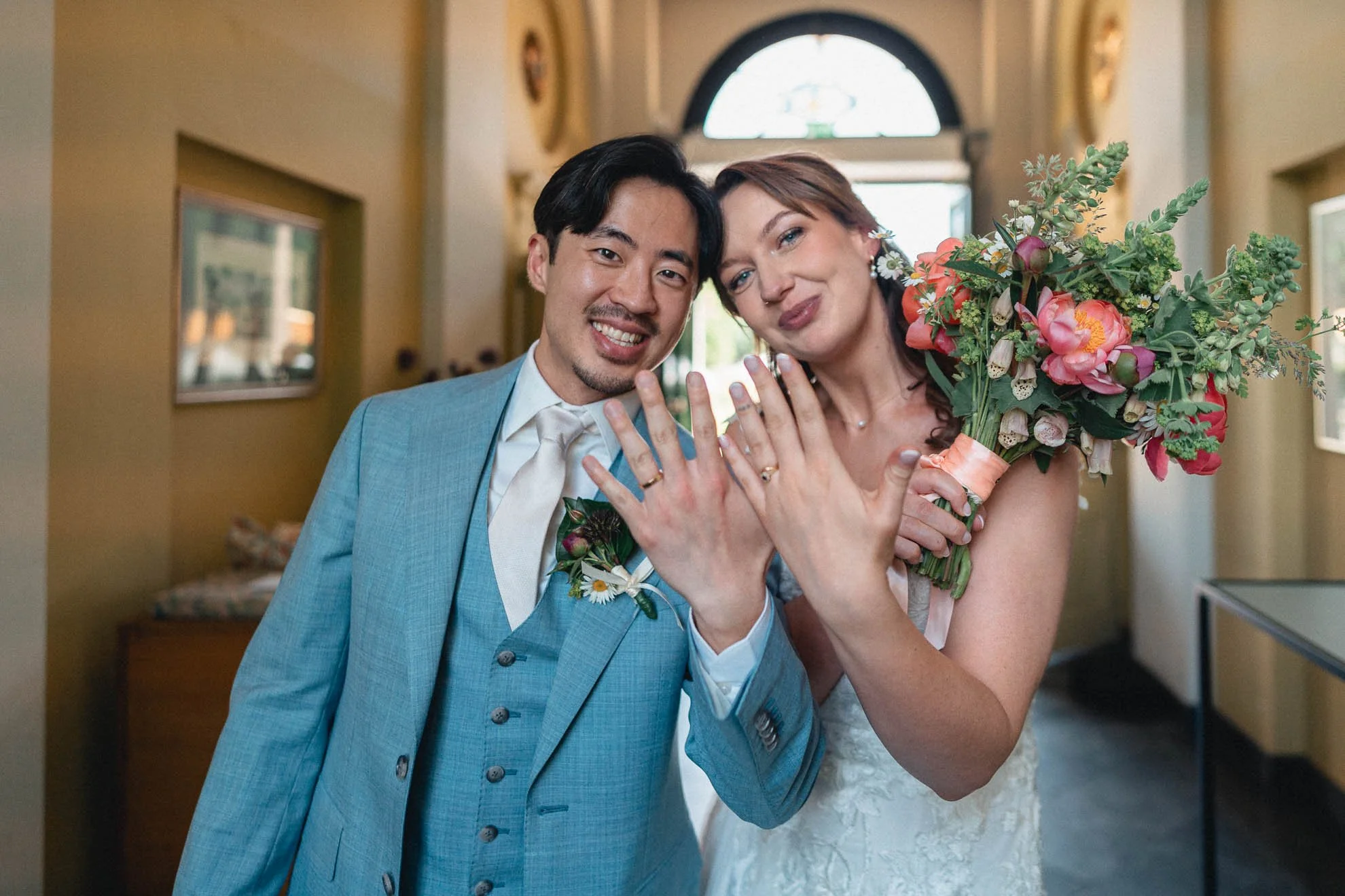 A happy man in a blue suit and a woman in a wedding dress are displaying their wedding rings while smiling and holding a bouquet of flowers. They are in a well-lit hallway with arched windows.