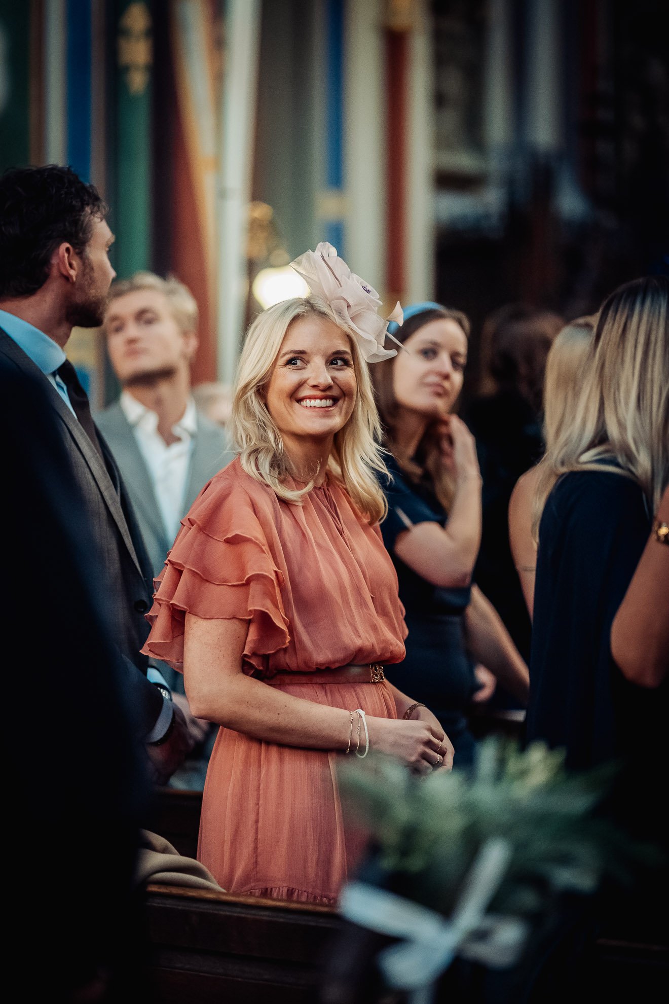 Woman with blonde hair smiling at an indoor event, wearing a peach-colored dress with ruffled sleeves and a large fascinator hat.