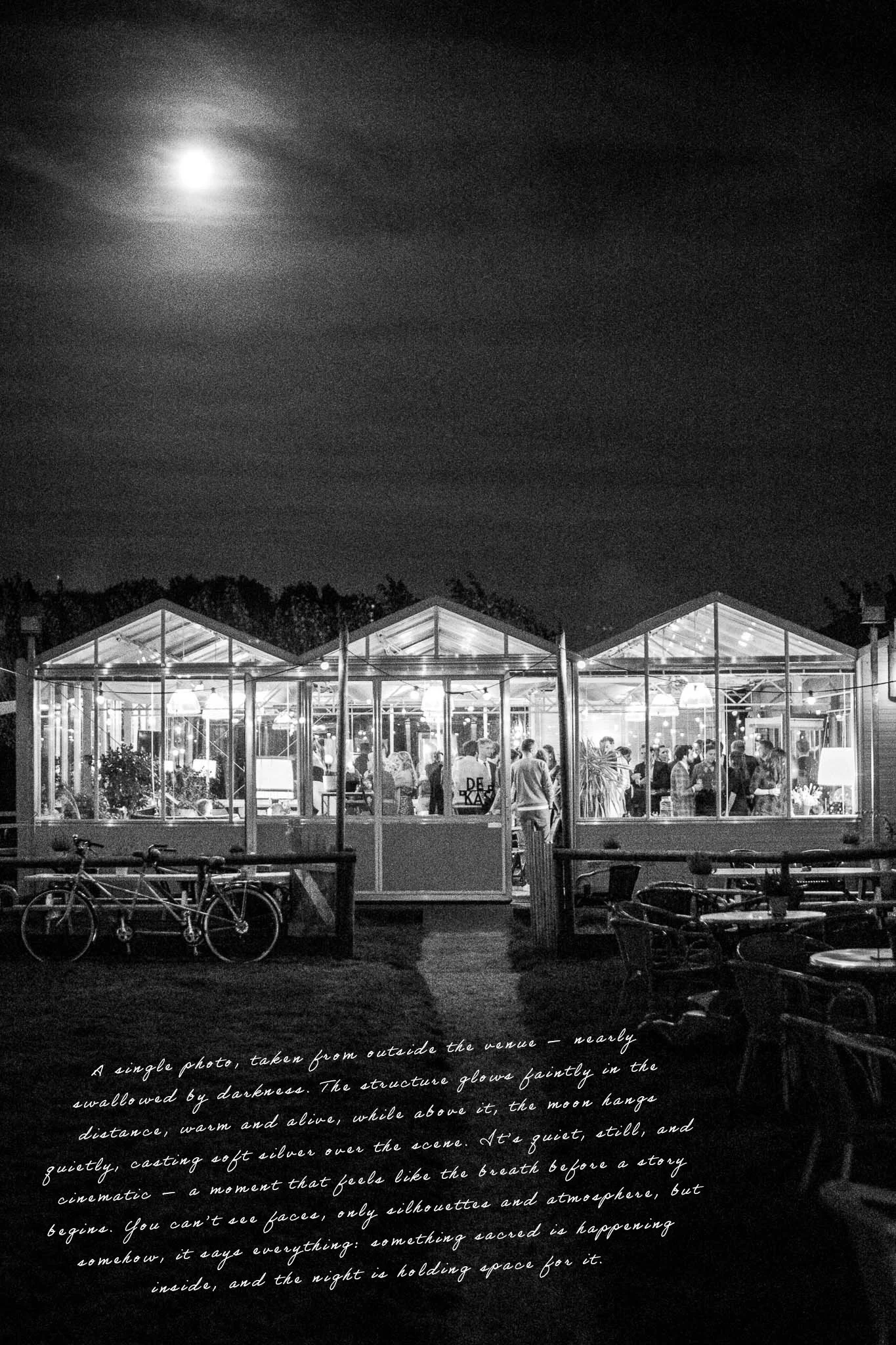 Een zwart-wit foto van een glazen gebouw met mensen die binnen staan. De maan schijnt in de donkere nacht boven het gebouw. Er staan fietsen en buiten tafels met stoelen.