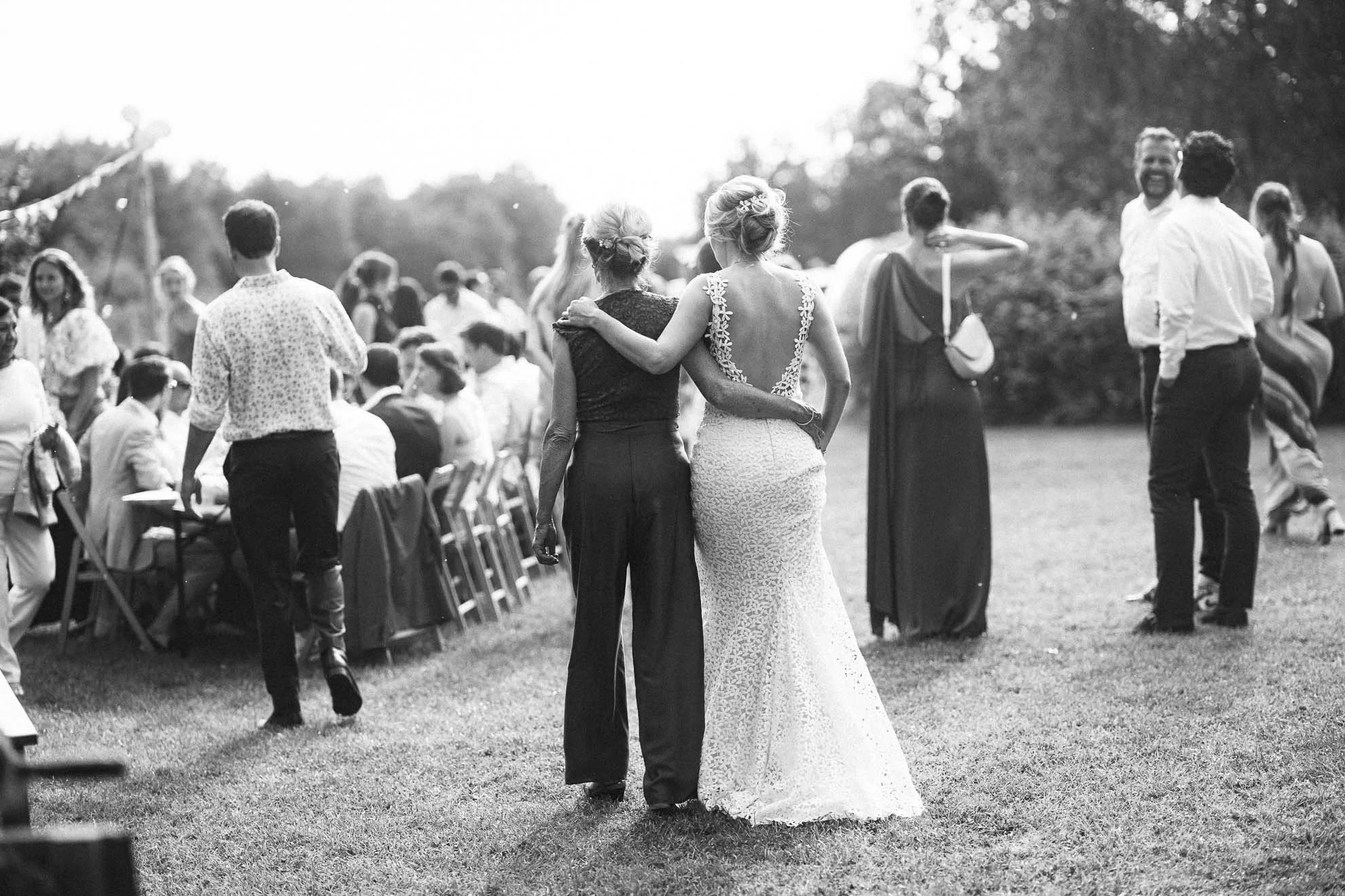 Black and white photo of a bride and her mother walking away from the camera at an outdoor wedding reception. The bride is wearing a lace wedding gown, and the mother is dressed in a dark outfit. People are seated and standing in the background, cele