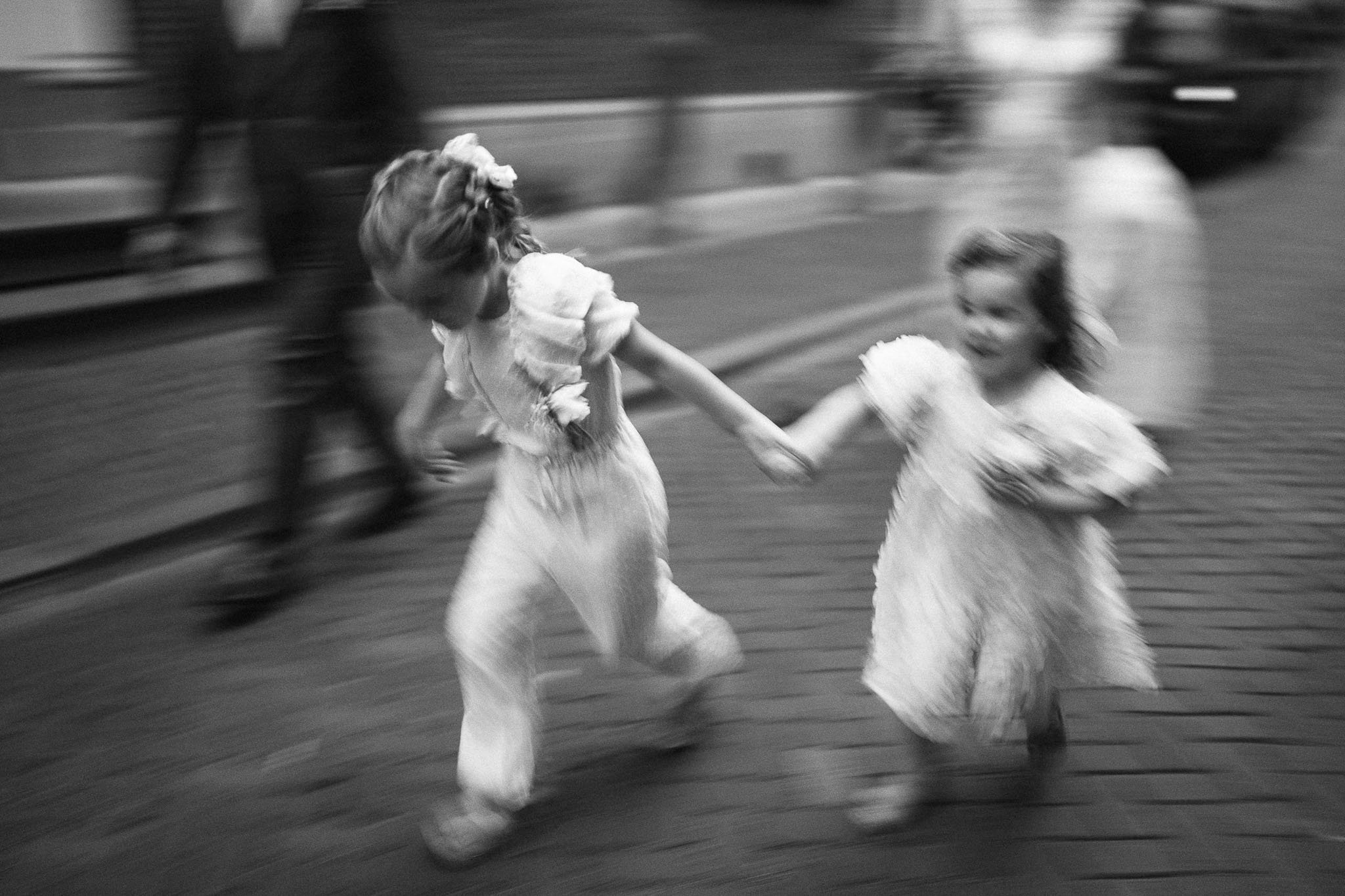 Twee jonge meisjes in witte jurkjes dansen samen op een straat, bewegend snel en lachend.