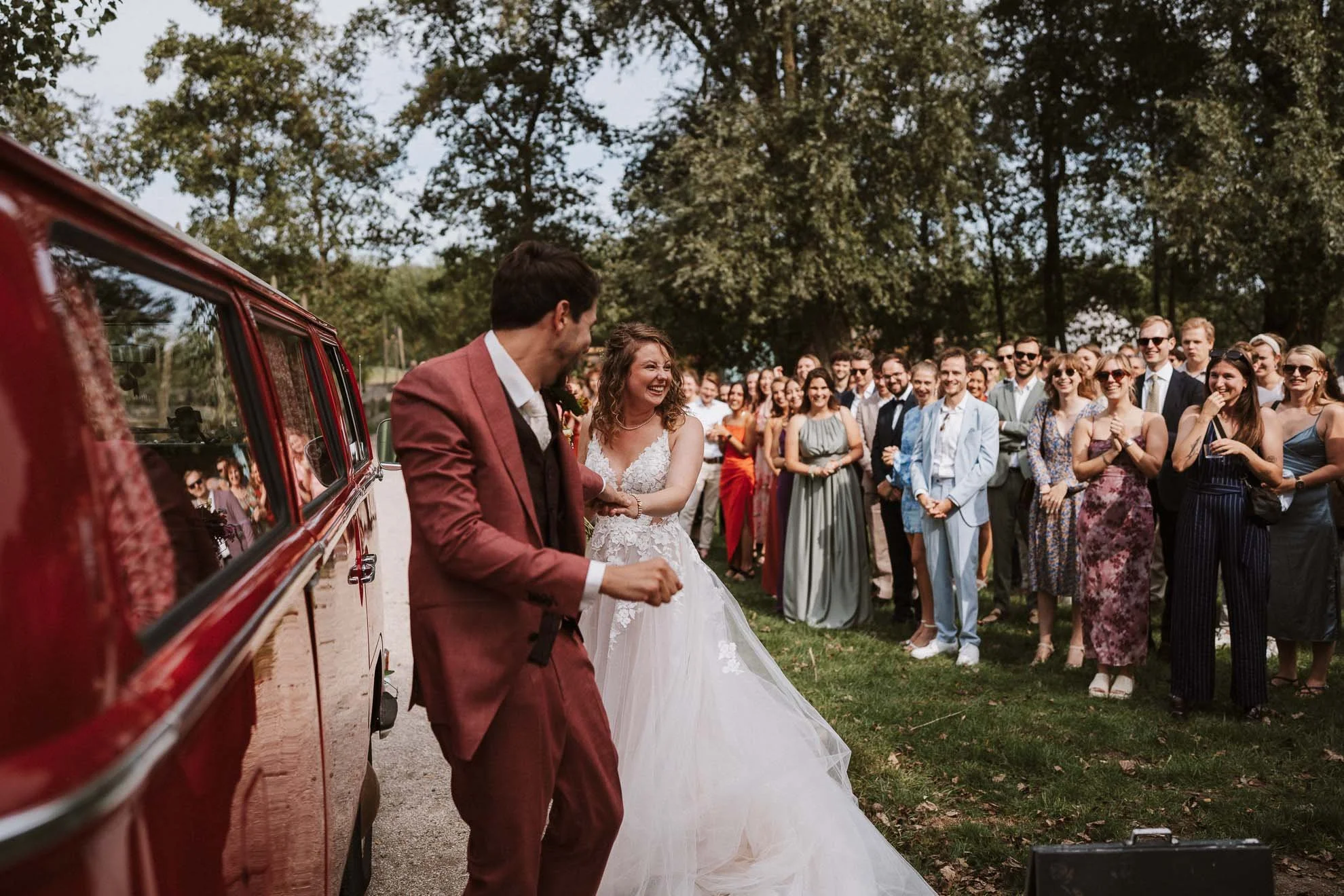 A bride and groom holding hands and smiling at each other outside in front of a crowd of wedding guests, with a red vintage car partially visible to the left, and trees in the background.