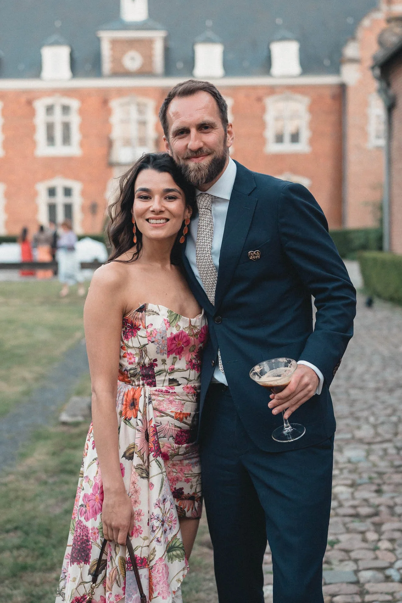A smiling woman and man dressed in formal attire standing outdoors in front of a large brick building with white window frames, holding drinks at what appears to be an outdoor event.