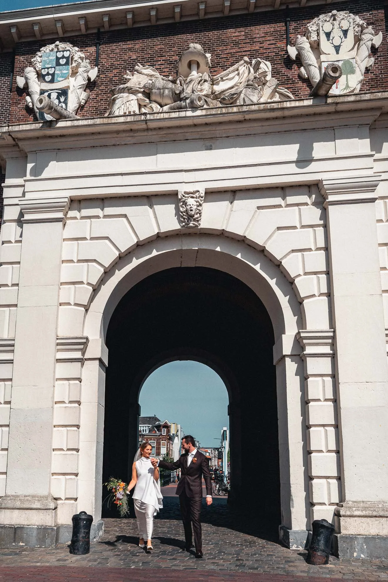 A bride and groom are walking under a large carved stone archway, with the bride wearing white and holding a bouquet, and the groom in a dark suit, in an urban setting with buildings in the background.