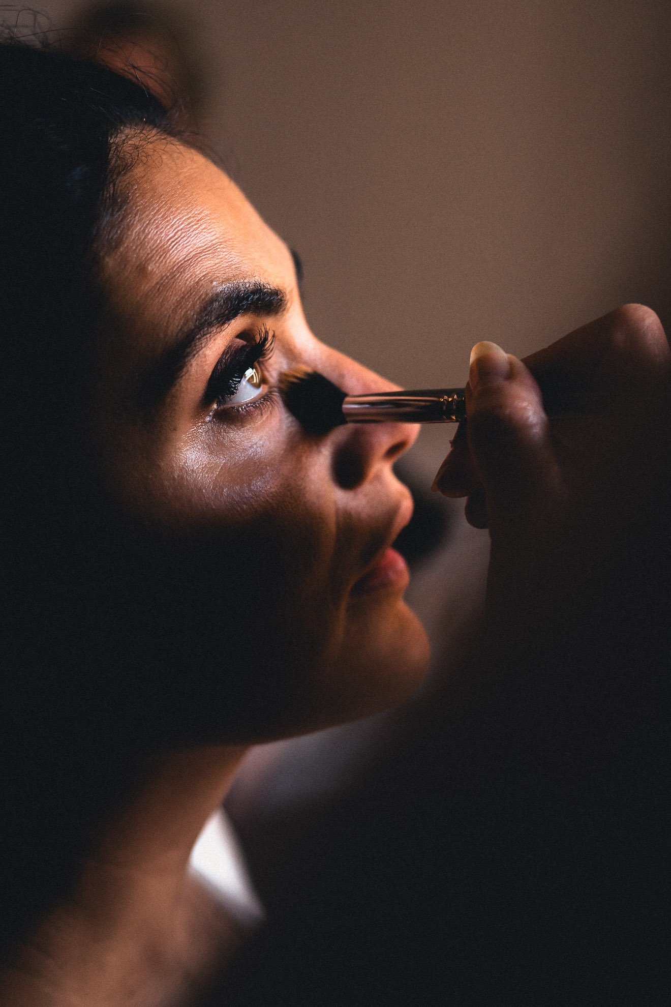 Close-up of a woman with dark hair and brown skin getting makeup applied with a brush near her nose, in a dimly lit setting.