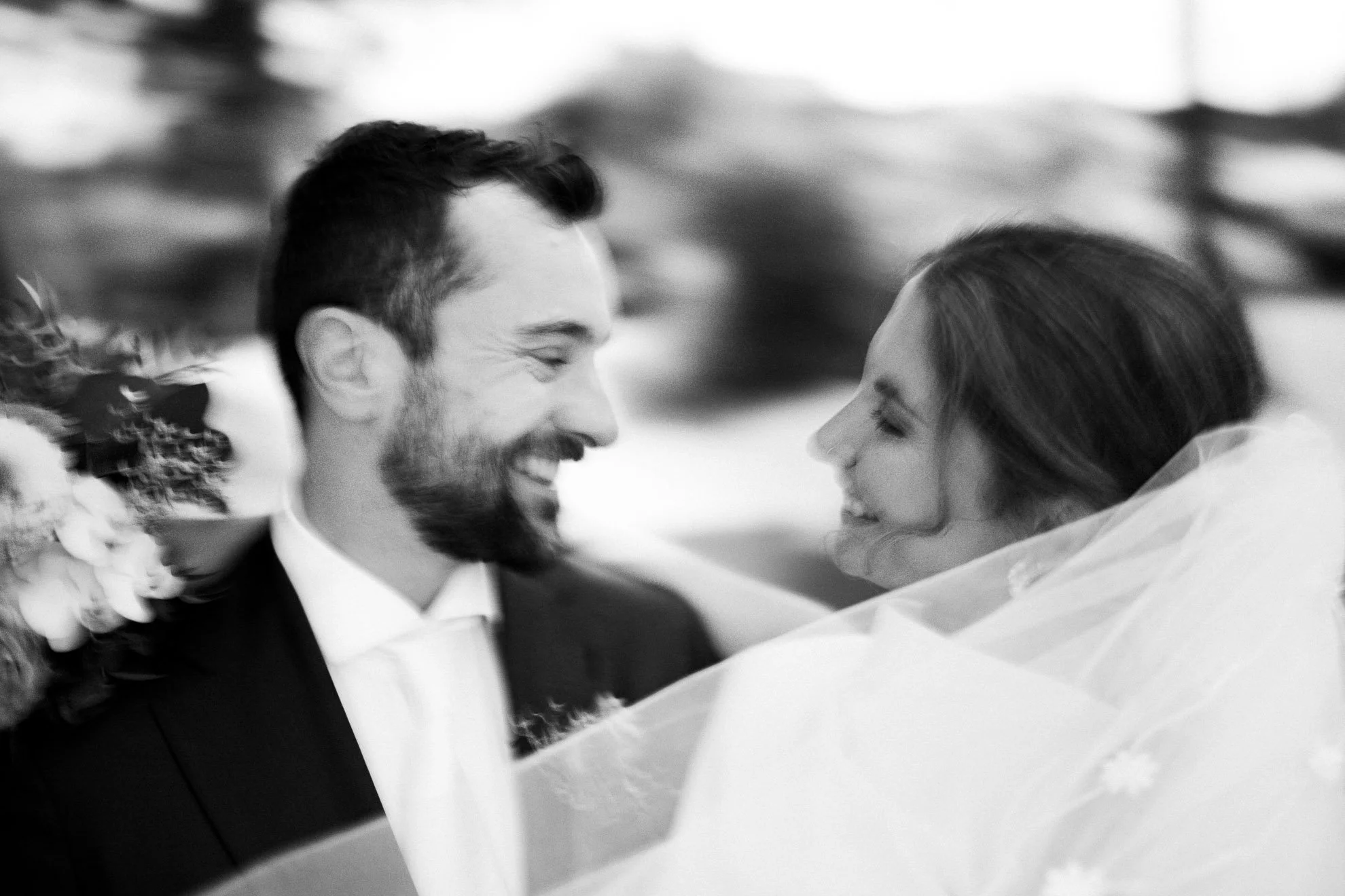 A black and white photo of a smiling man and woman close together, possibly at a wedding or celebration, with the man wearing a tuxedo and the woman in a wedding dress.