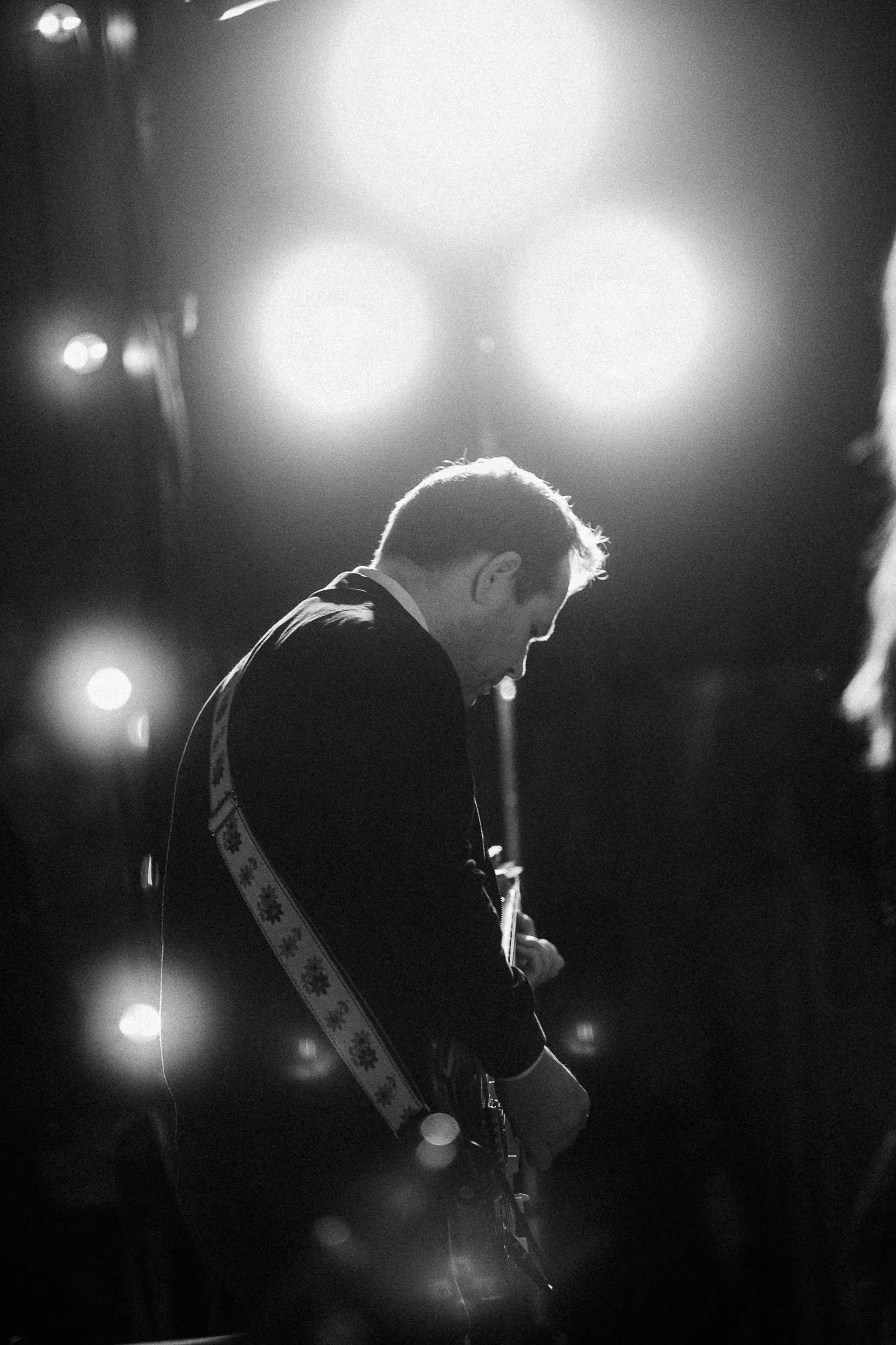 A black and white photo of a person playing guitar or ukulele, with lights in the background creating a bokeh effect. The person's face is turned downward, and they appear to be on stage or in a performance setting.