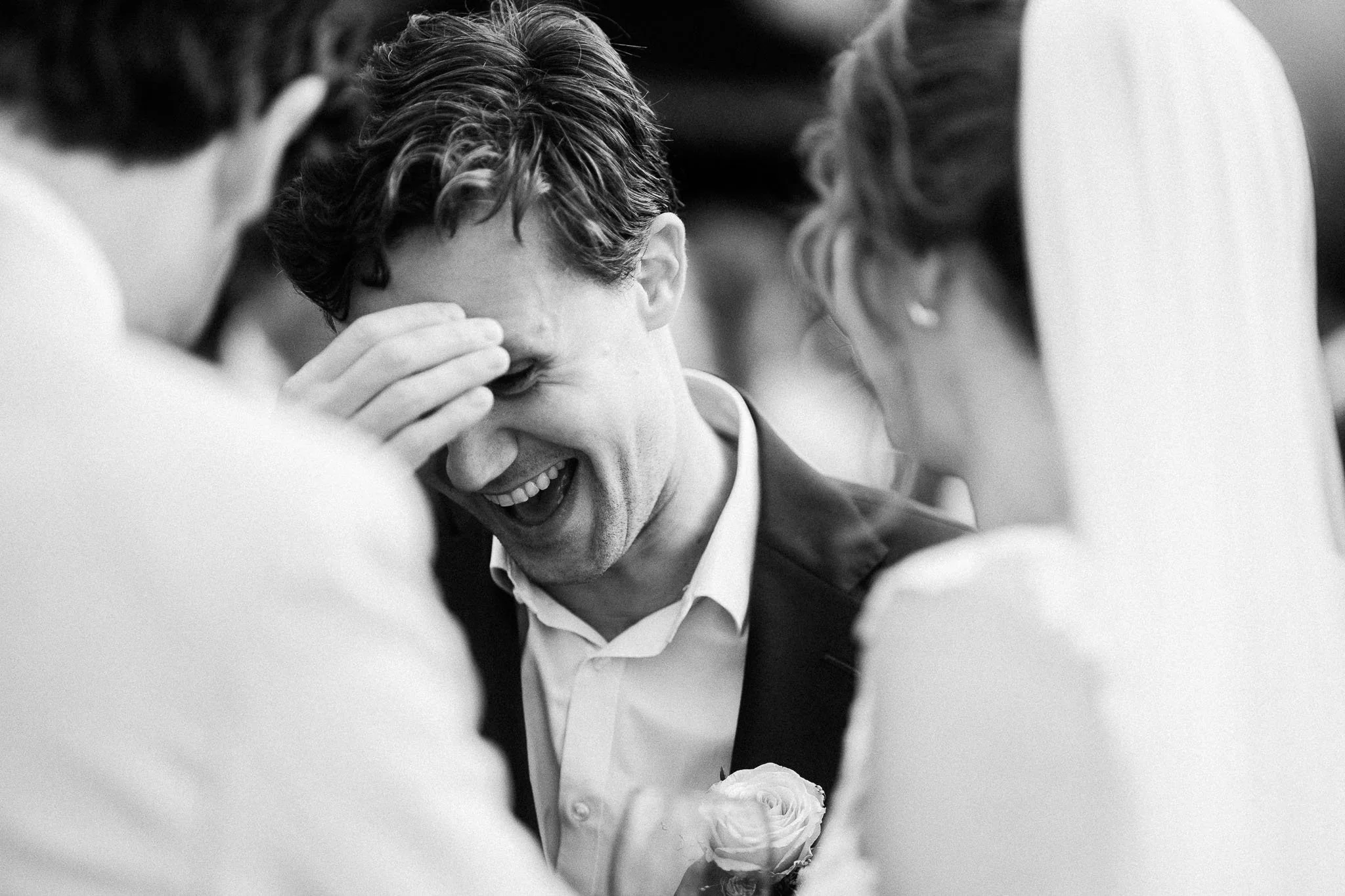 Een man lacht met gesloten ogen terwijl hij omringd wordt door twee mensen, een vrouw en een man, op een feest of bruiloft. De man heeft een witte bloem op zijn jas en lijkt een emotionele of gelukkige moment te beleven.