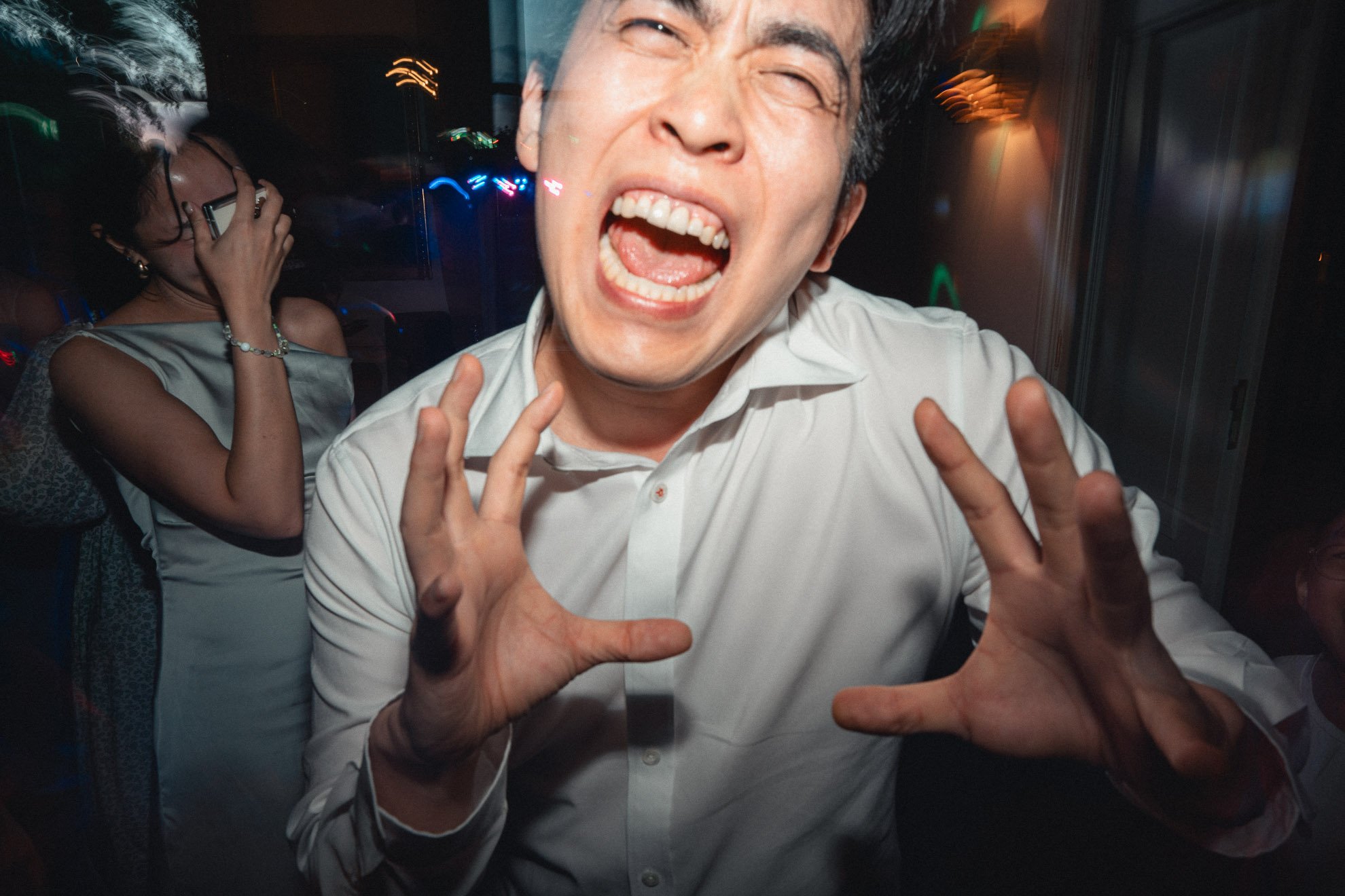 A man with gray hair and a white shirt smiling and making a framing gesture with his fingers at the camera in a party or nightclub setting.