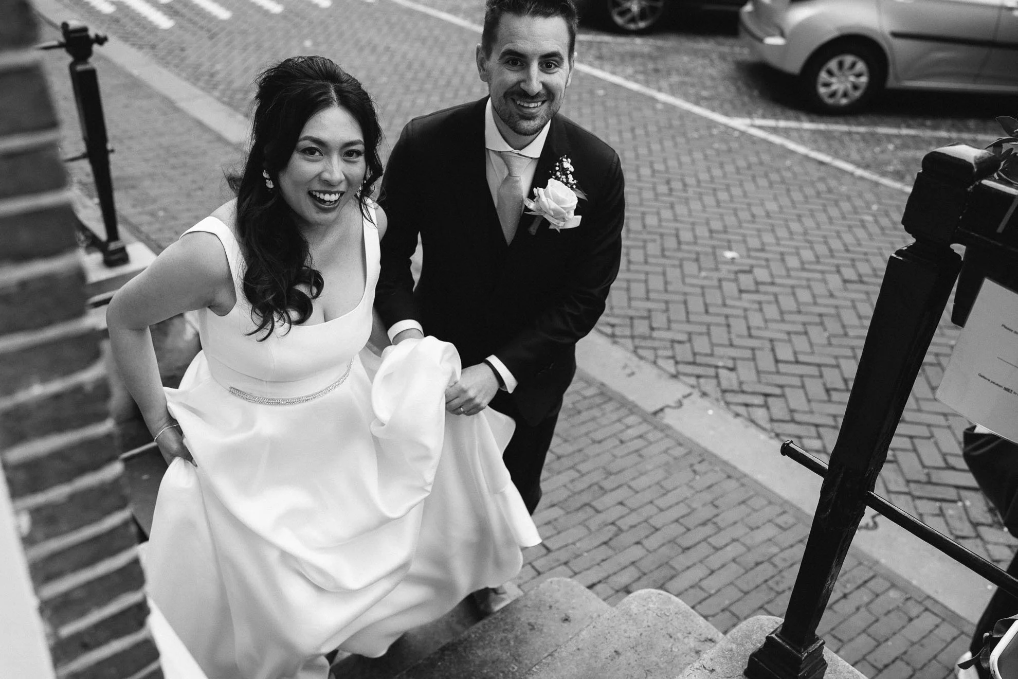 A smiling couple at a wedding, standing on a brick sidewalk, with vehicles in the background. The bride is wearing a white wedding dress, and the groom is in a dark suit with a boutonniere. The photo is in black and white.