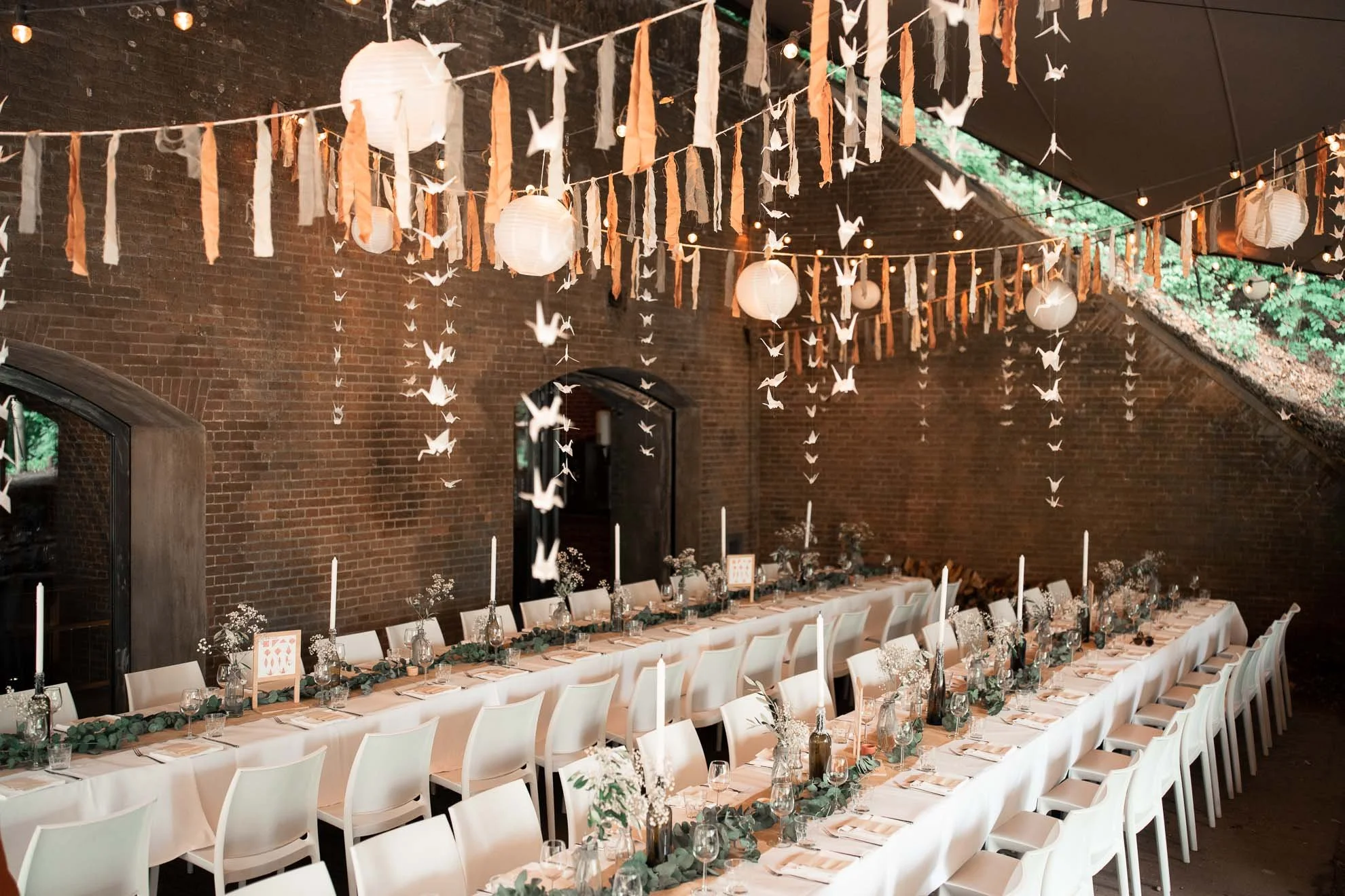 Decorative string lights, paper lanterns, origami cranes, and torn fabric banners hanging from the ceiling of a rustic event space with a long banquet table decorated with candles, flowers, and greenery.