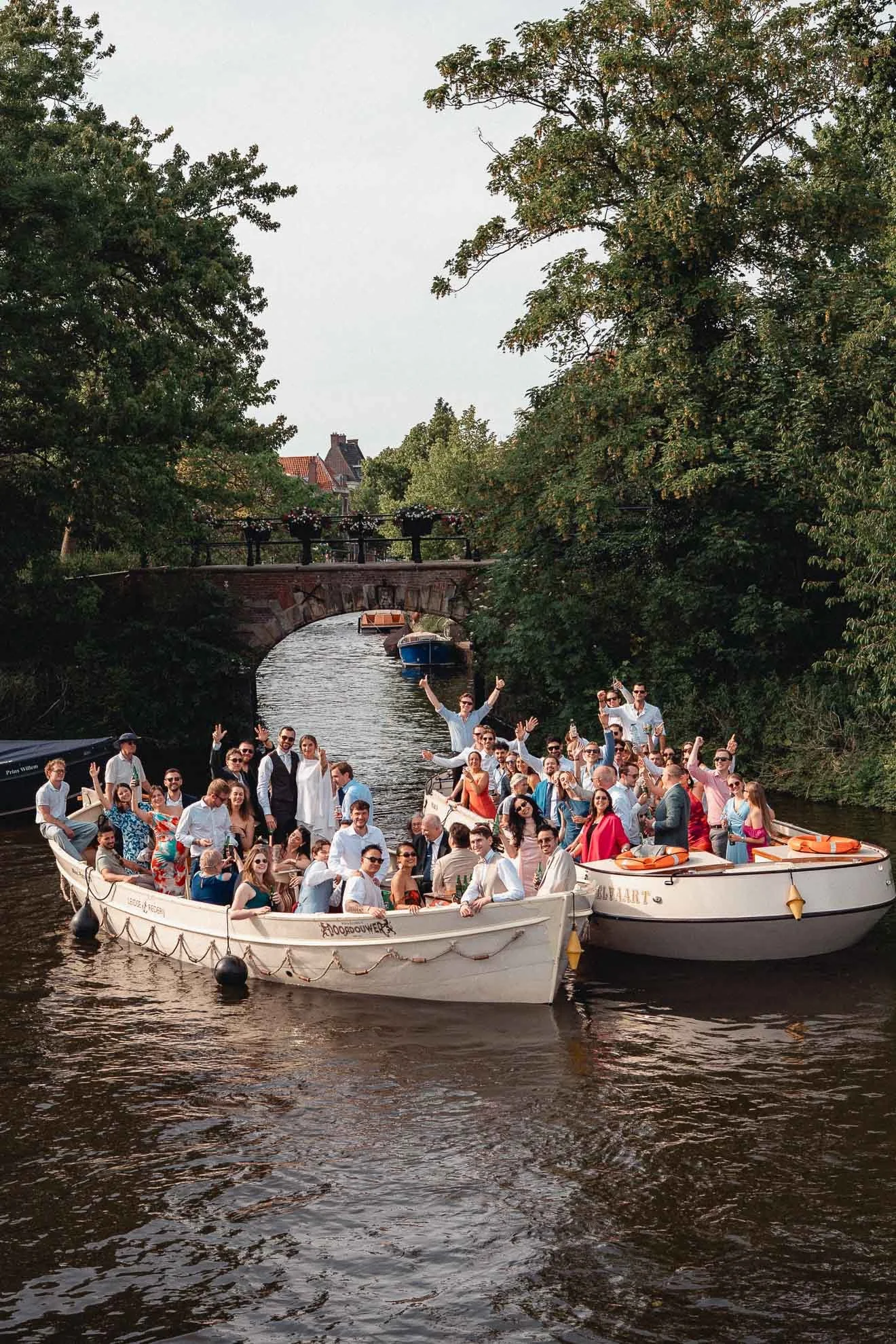 A large group of people celebrating on a boat decorated for a special occasion, on a river with trees and a bridge in the background.